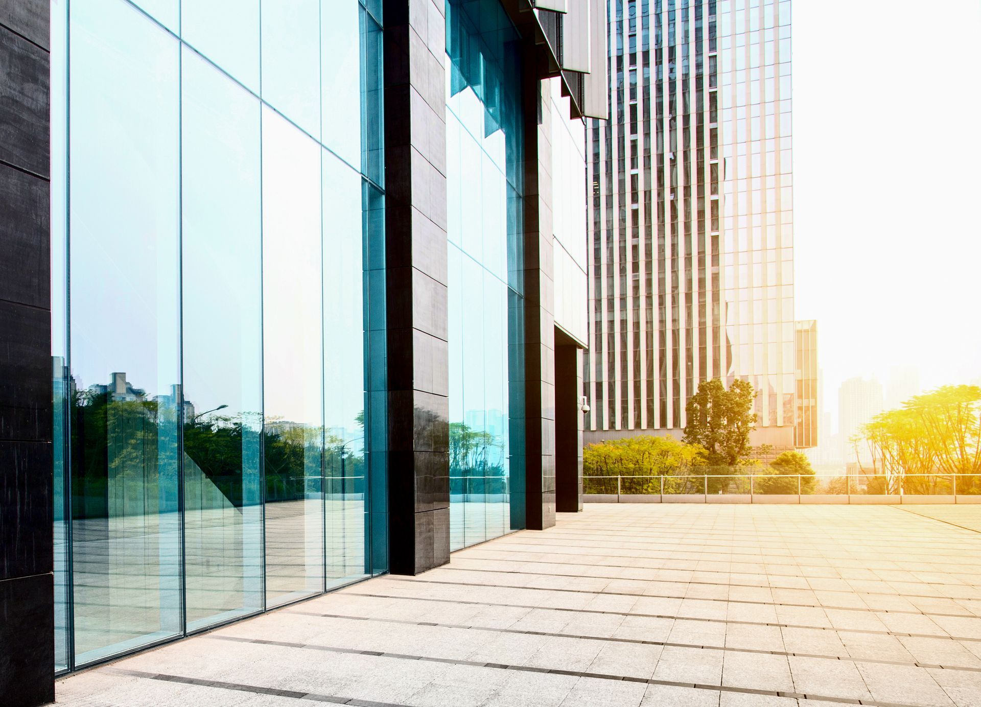 Modern building with glass walls and a tiled walkway, another skyscraper in background. Bright sunlight.