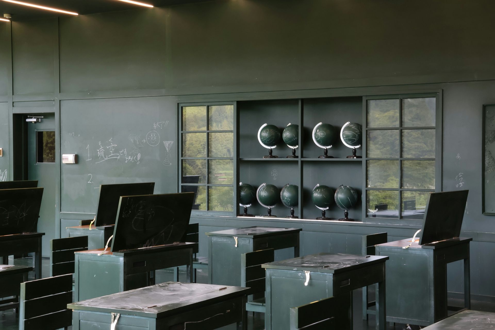 Empty, dark green classroom with desks, globes, and windows; likely an old school.
