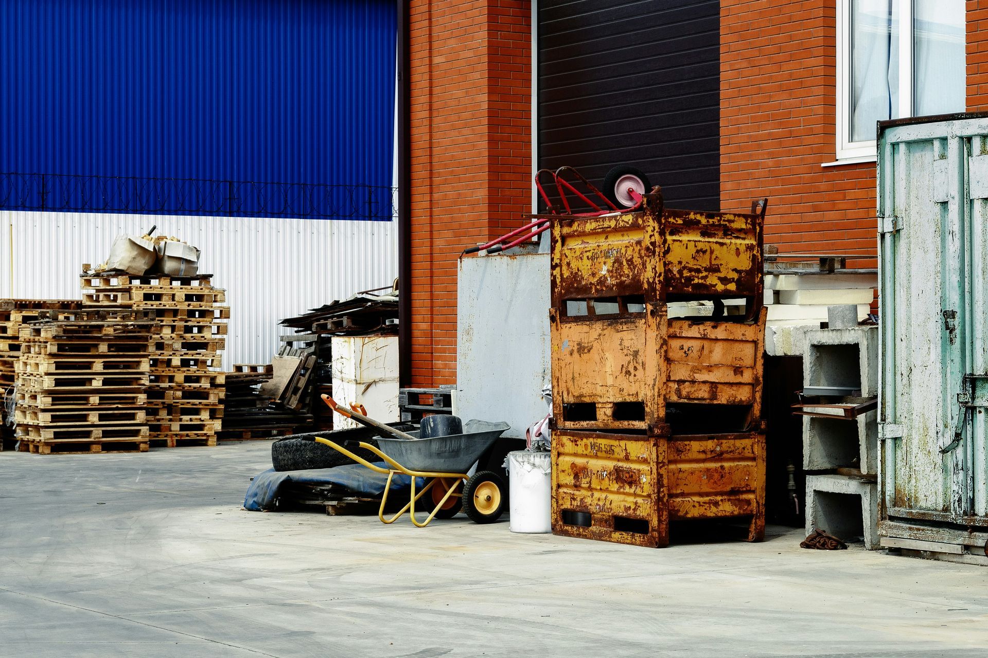 Exterior of a warehouse with stacked pallets, rusty metal containers, and brick and blue walls.