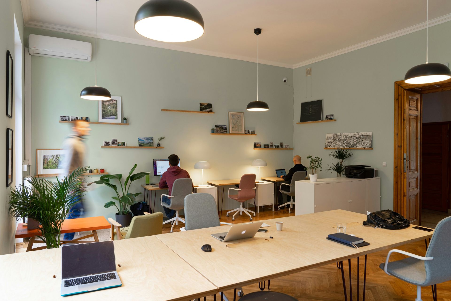 Office space with multiple desks, three people working, green walls, potted plants, and hanging lights.