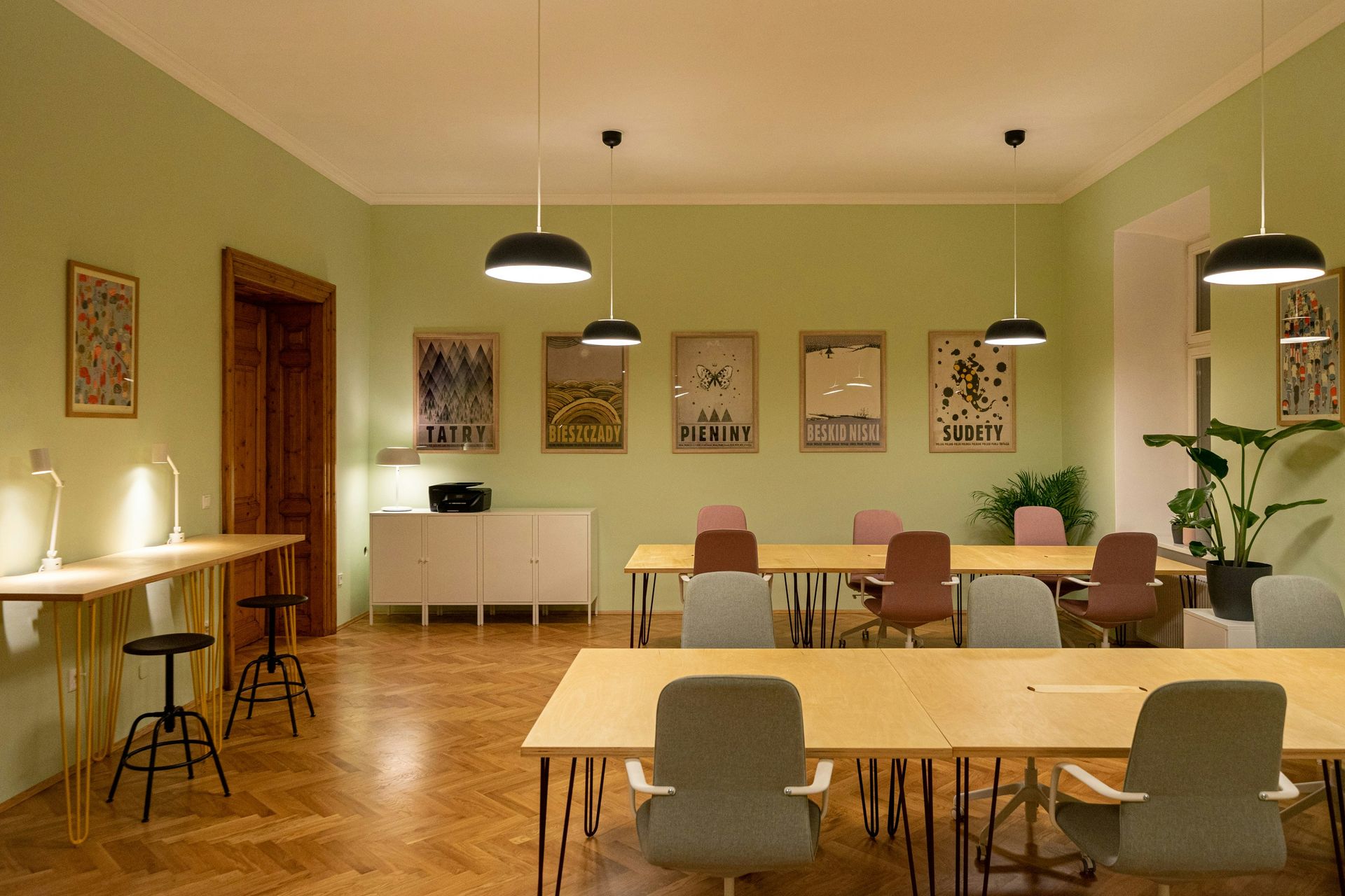 An office space with light green walls, wooden floors, tables, chairs, and artwork. Overhead lamps illuminate the room.
