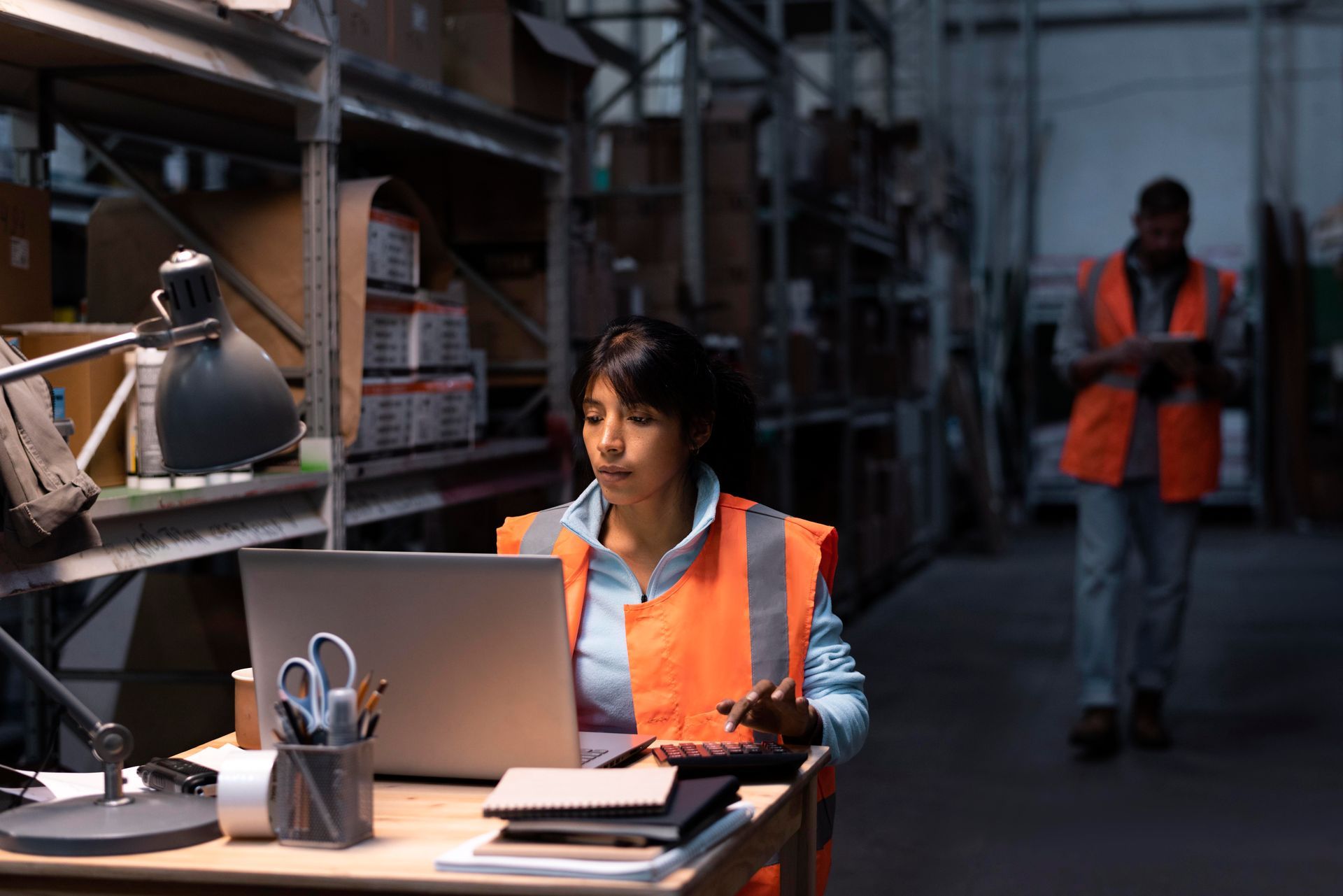 Woman in orange vest works on a laptop in a warehouse, another person walks in the background.