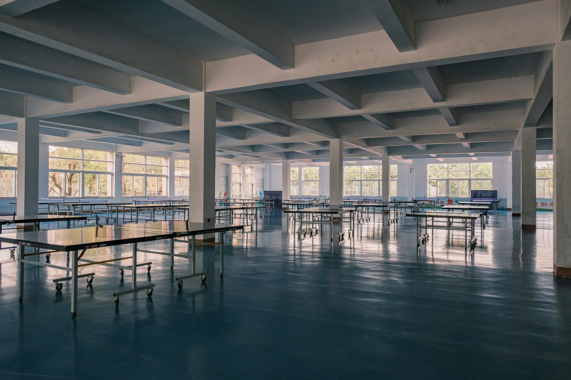 Empty cafeteria with blue floors, tables, white columns, and windows along walls.