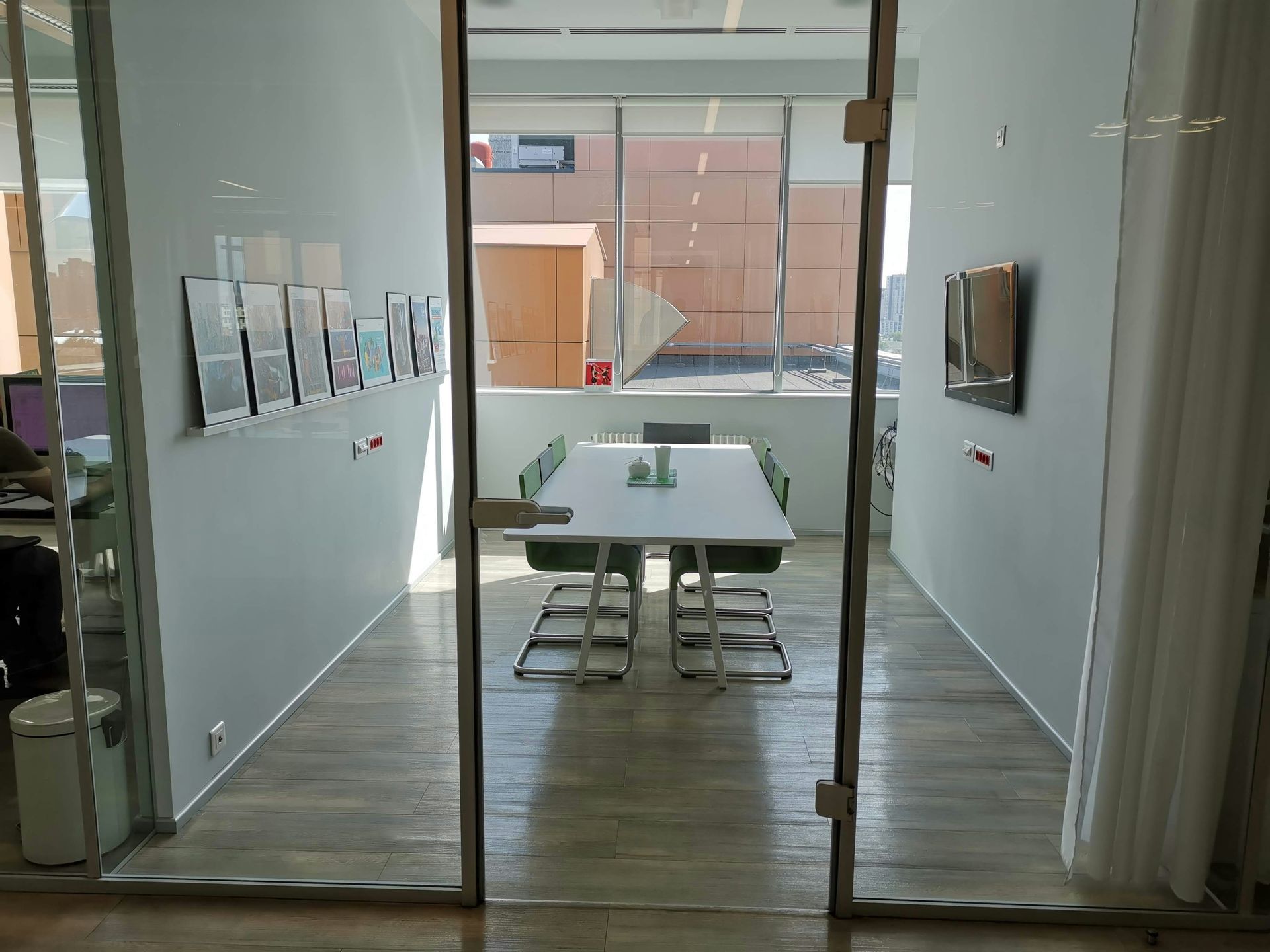 Meeting room through glass doors; white table, green chairs, light wood floor, posters on wall.