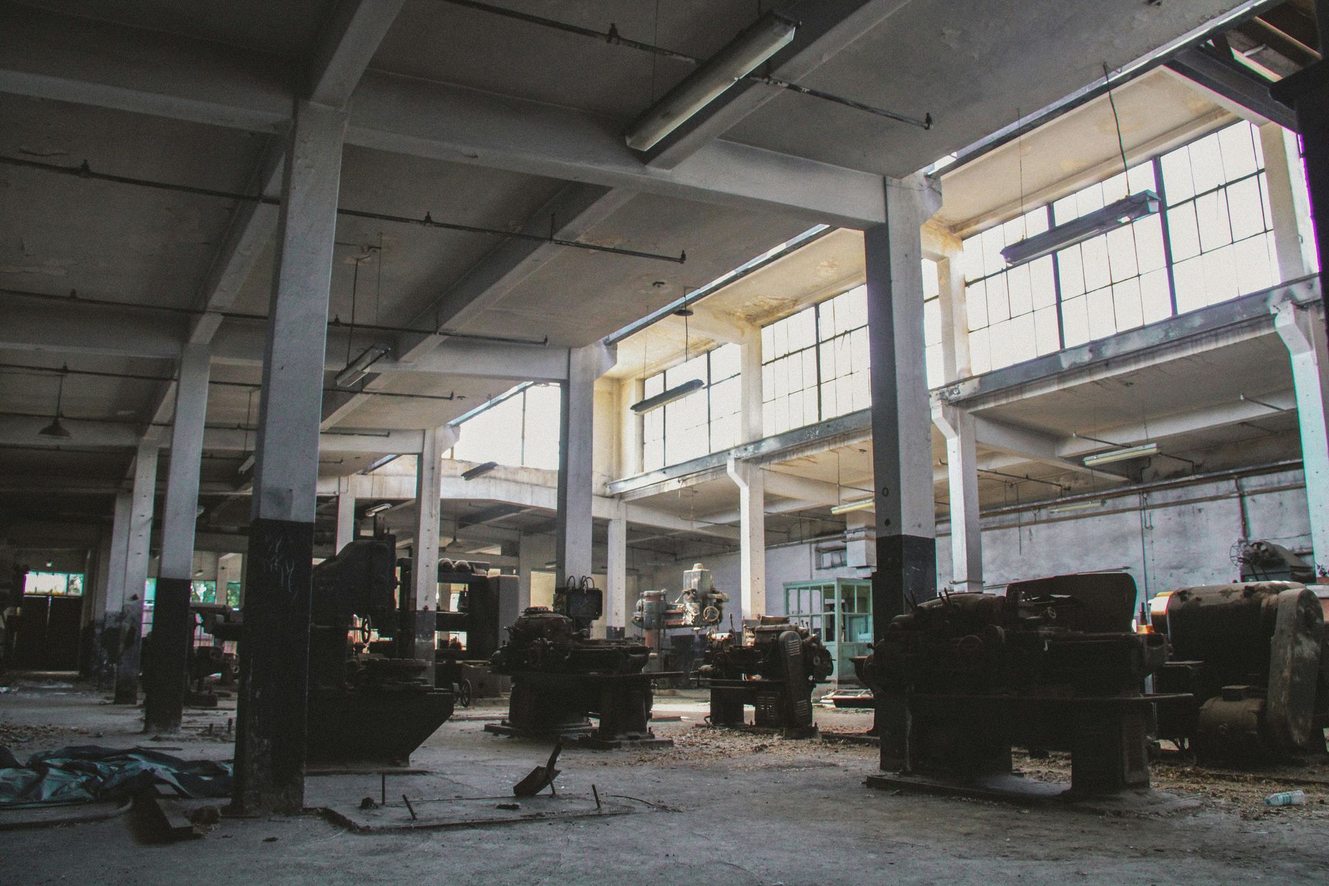 Interior of an abandoned factory with machinery, concrete pillars, and bright windows.