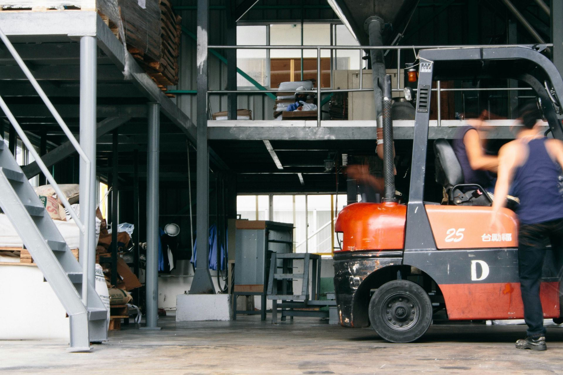 Forklift in warehouse. Person near the forklift, blurred motion. Metal structure, stairs, and windows in background.