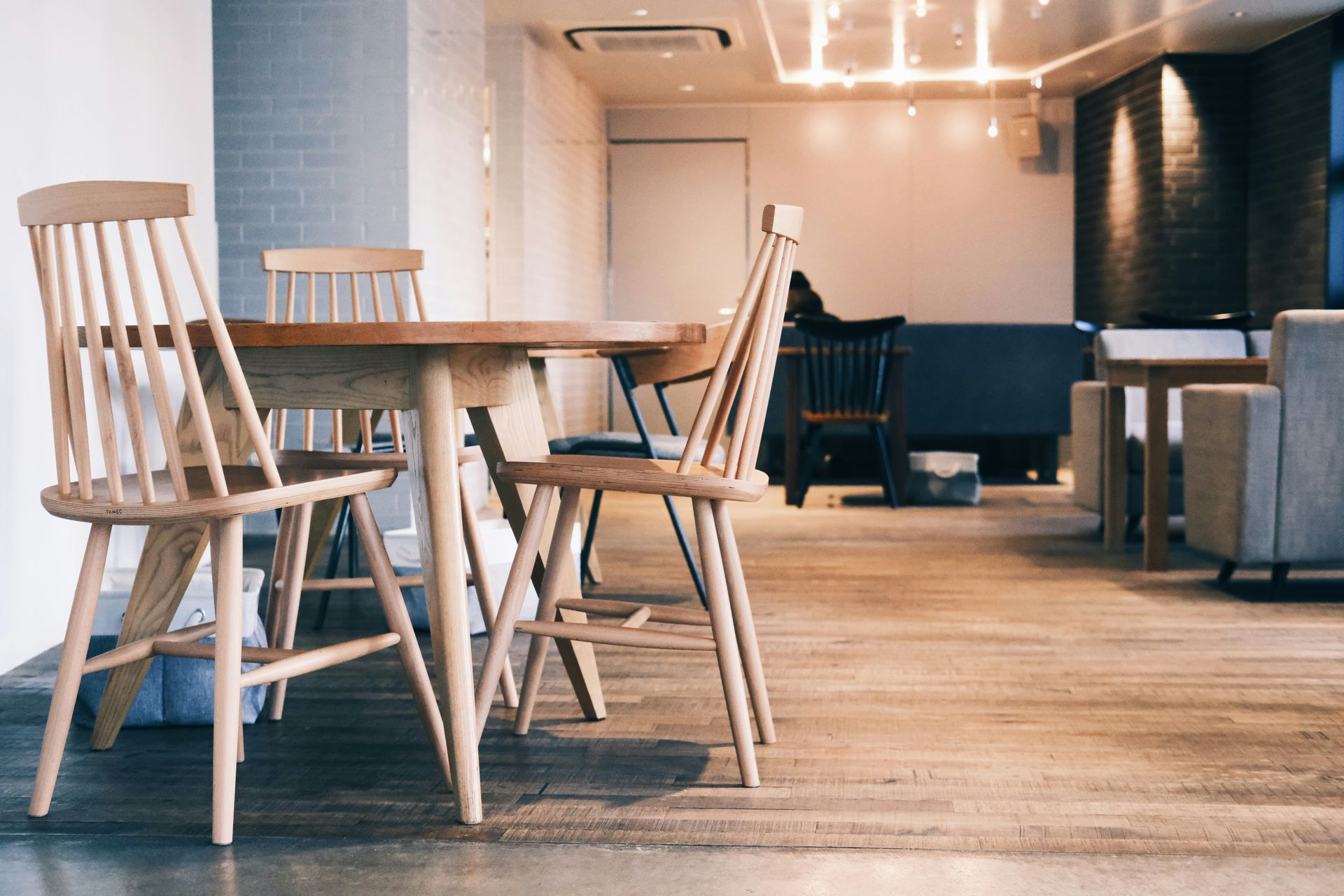 Wooden table and chairs in a cafe, neutral colors. Other seating and lighting in the background.