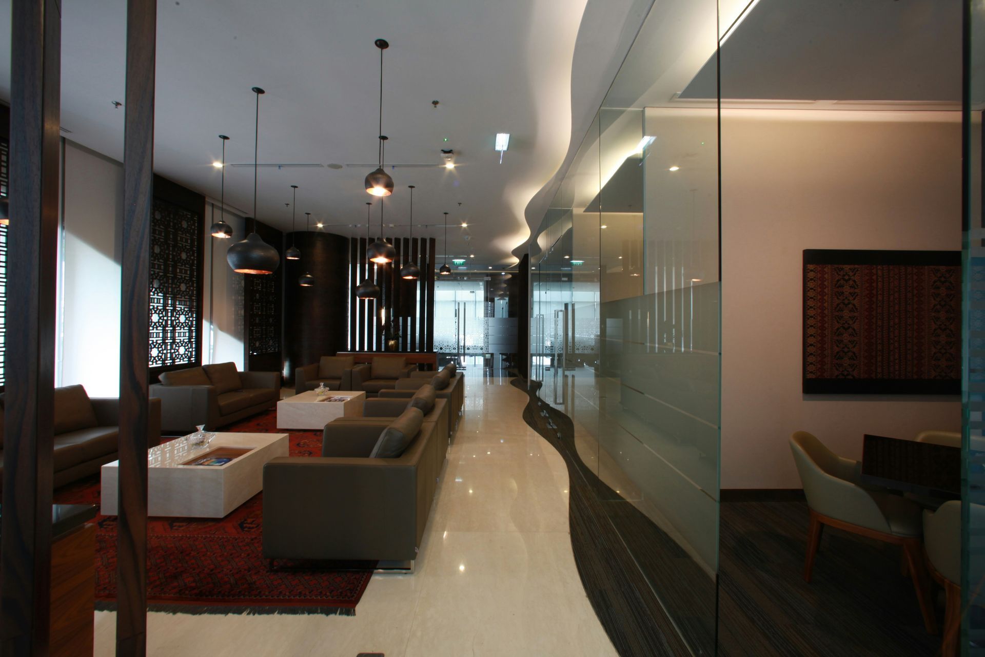 Lounge area with gray couches, white tables, and patterned rug. Modern design with glossy floors and glass walls.