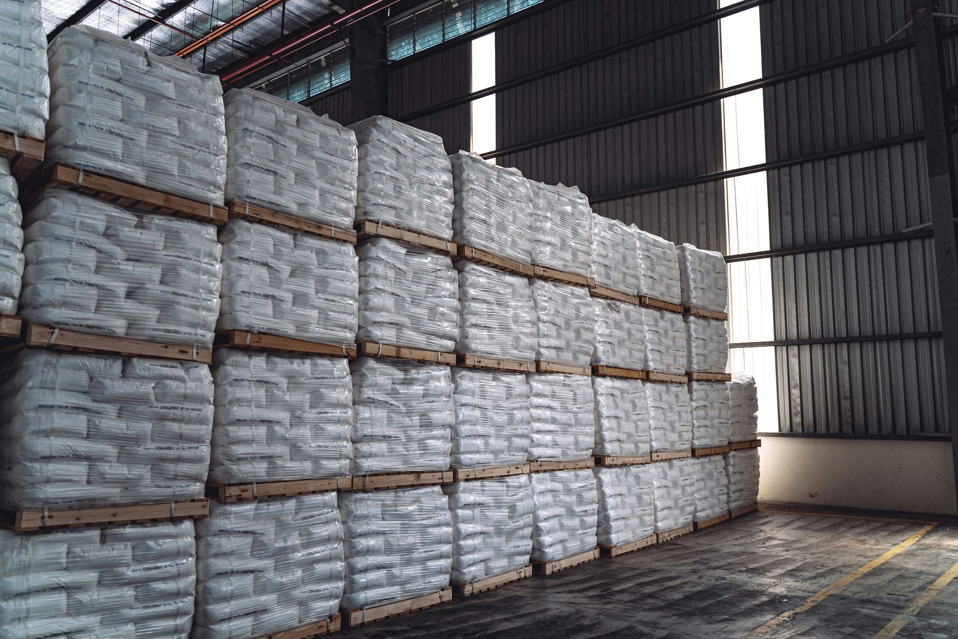 Stacks of white bags on wooden pallets inside a warehouse, illuminated by natural light.