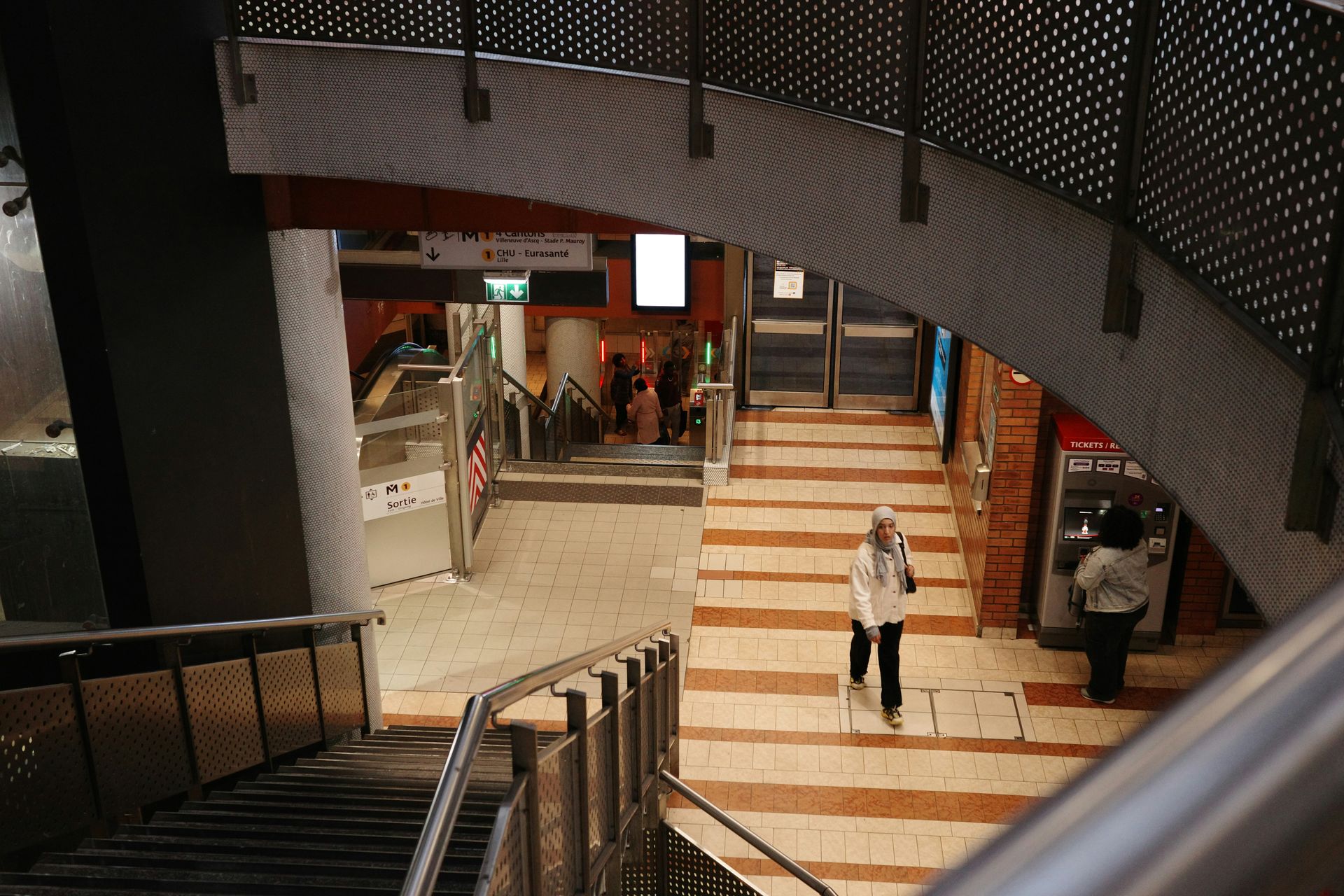 Staircase in a subway station with two people walking. Architecture features a metal railing and brick walls.