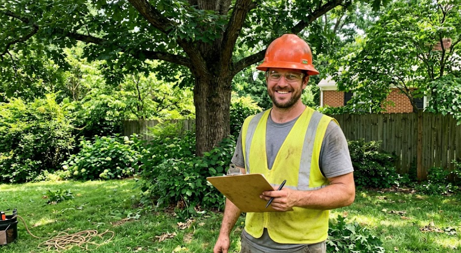 A professional arborist from Tree Service of Charlottesville smiling in a sunny backyard holding a clipboard.