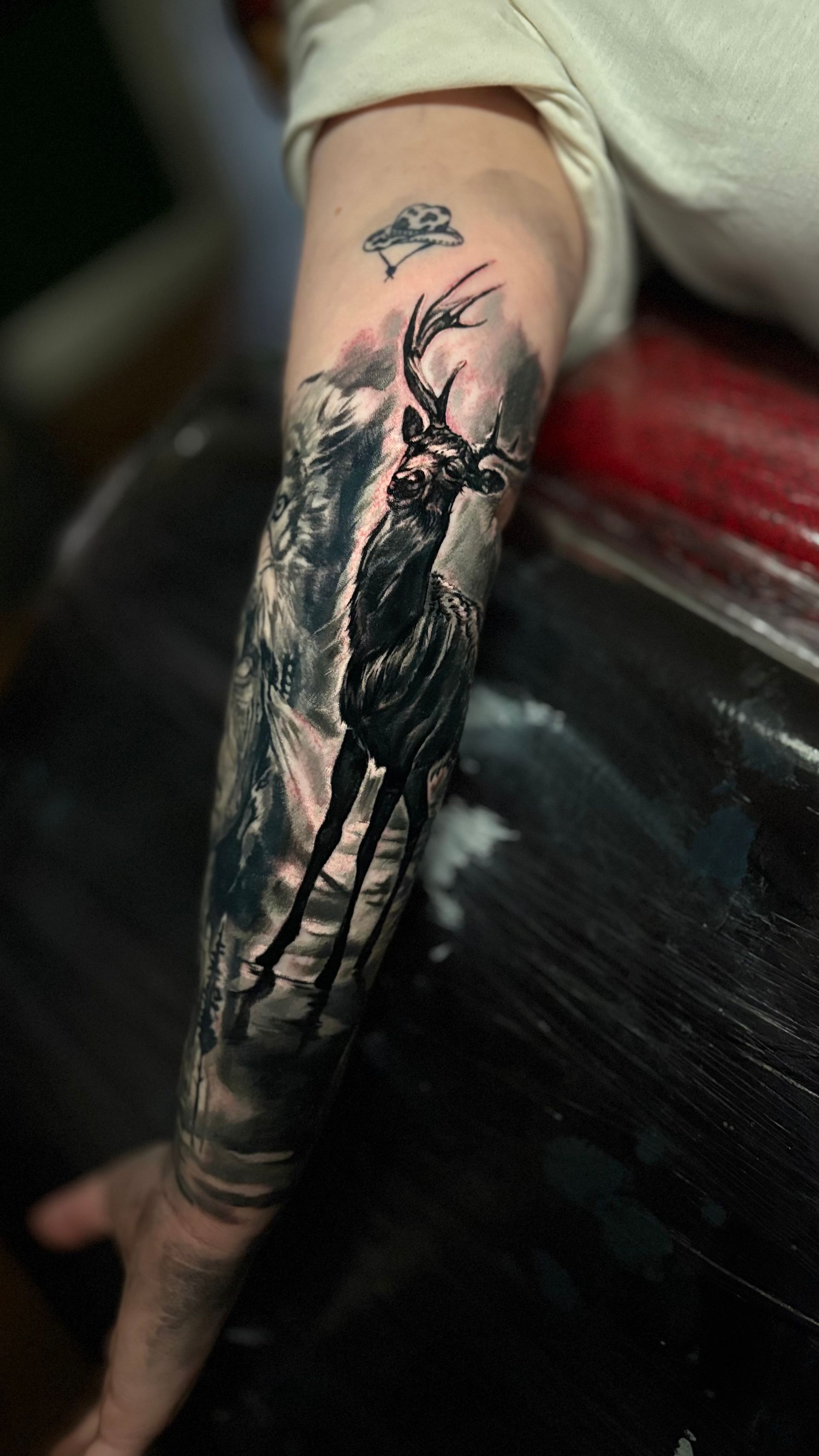 Arm tattoo of a stylized black deer with large antlers, set in a misty, dark forest scene.