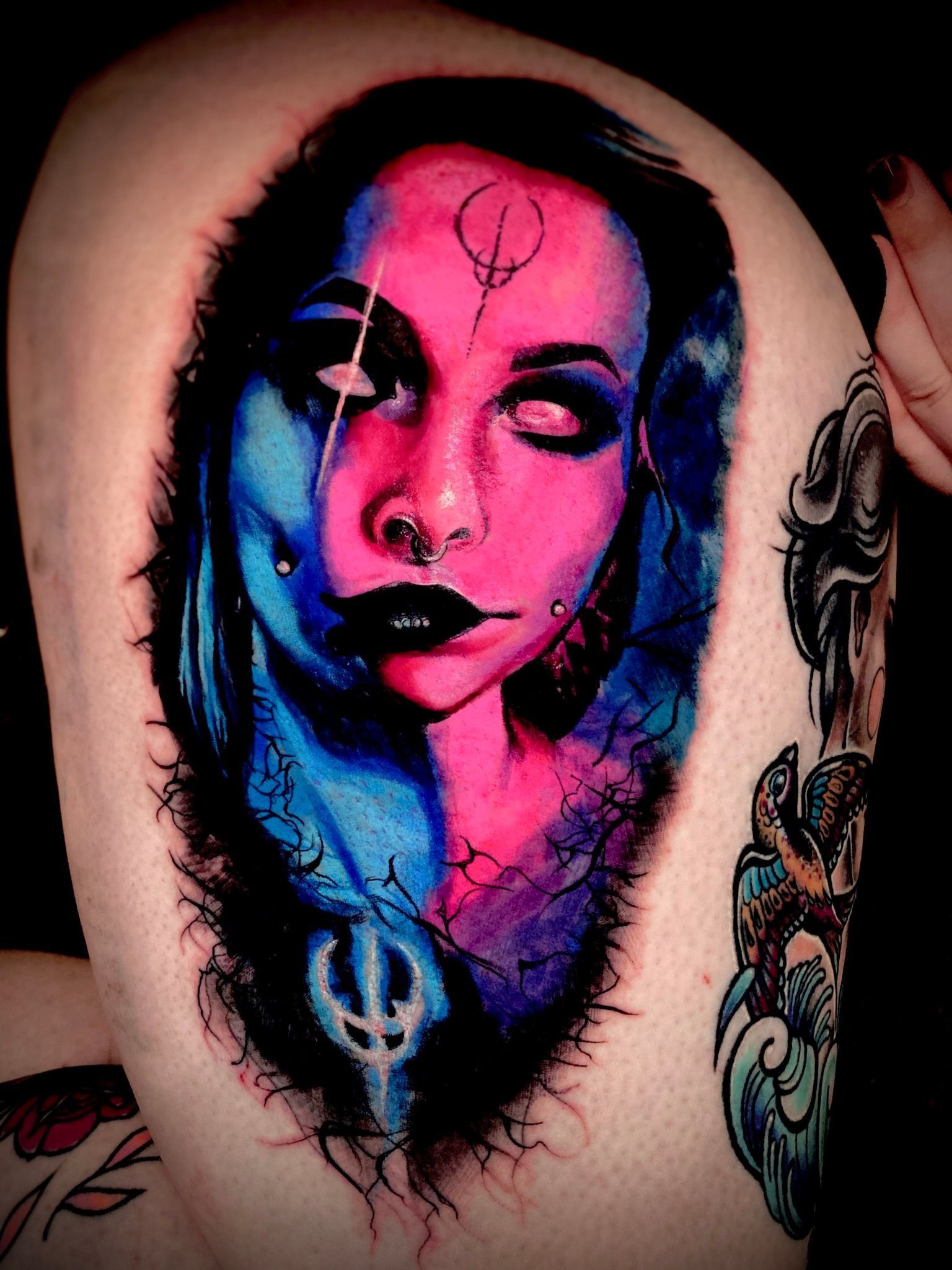 Tattoo on skin: split-face portrait, pink/blue color scheme, white eyes, dark lips, with symbols.