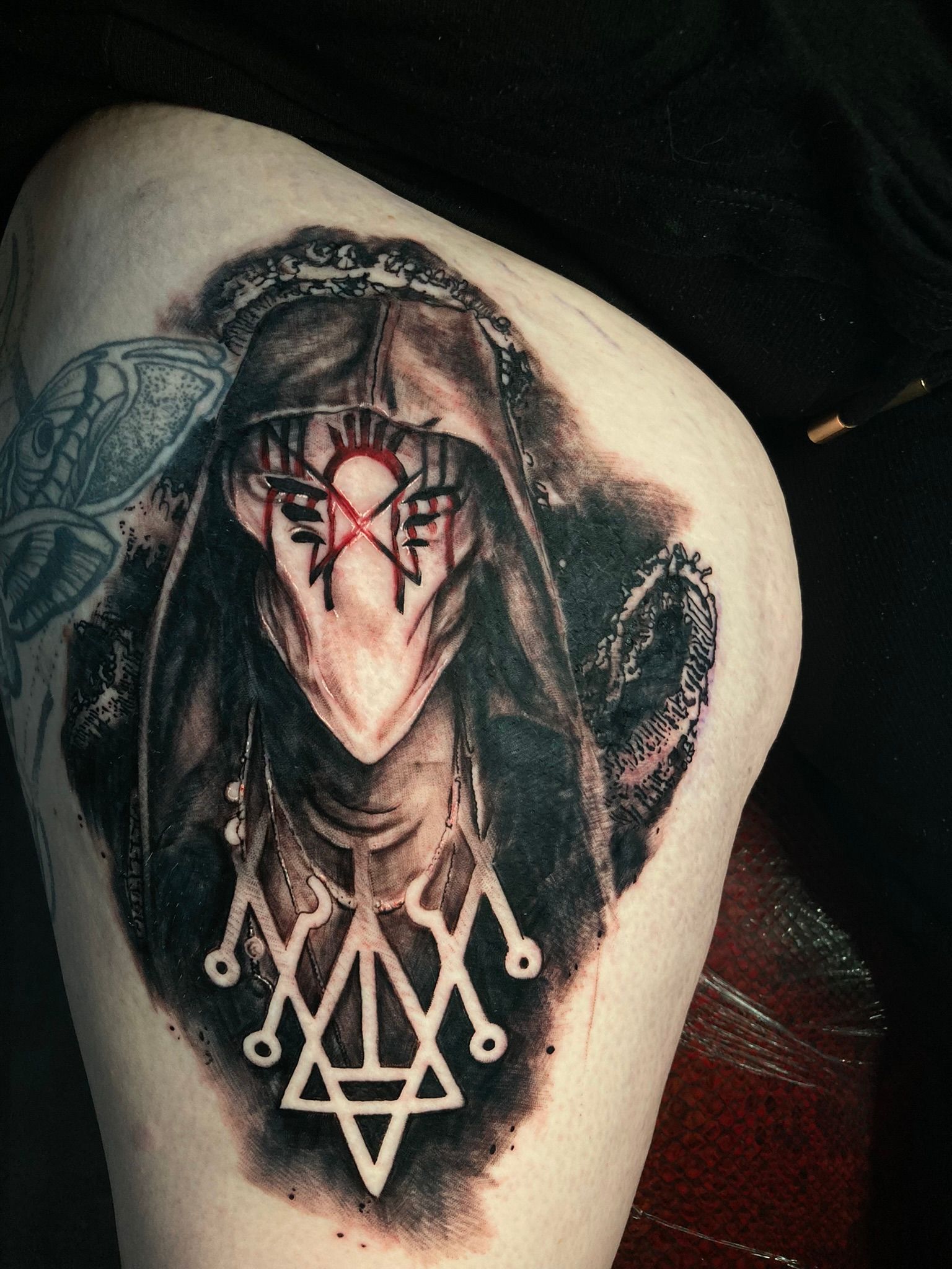 Tattoo of a hooded figure with a crown, mask, and a symbol below. Black and white with red accents.