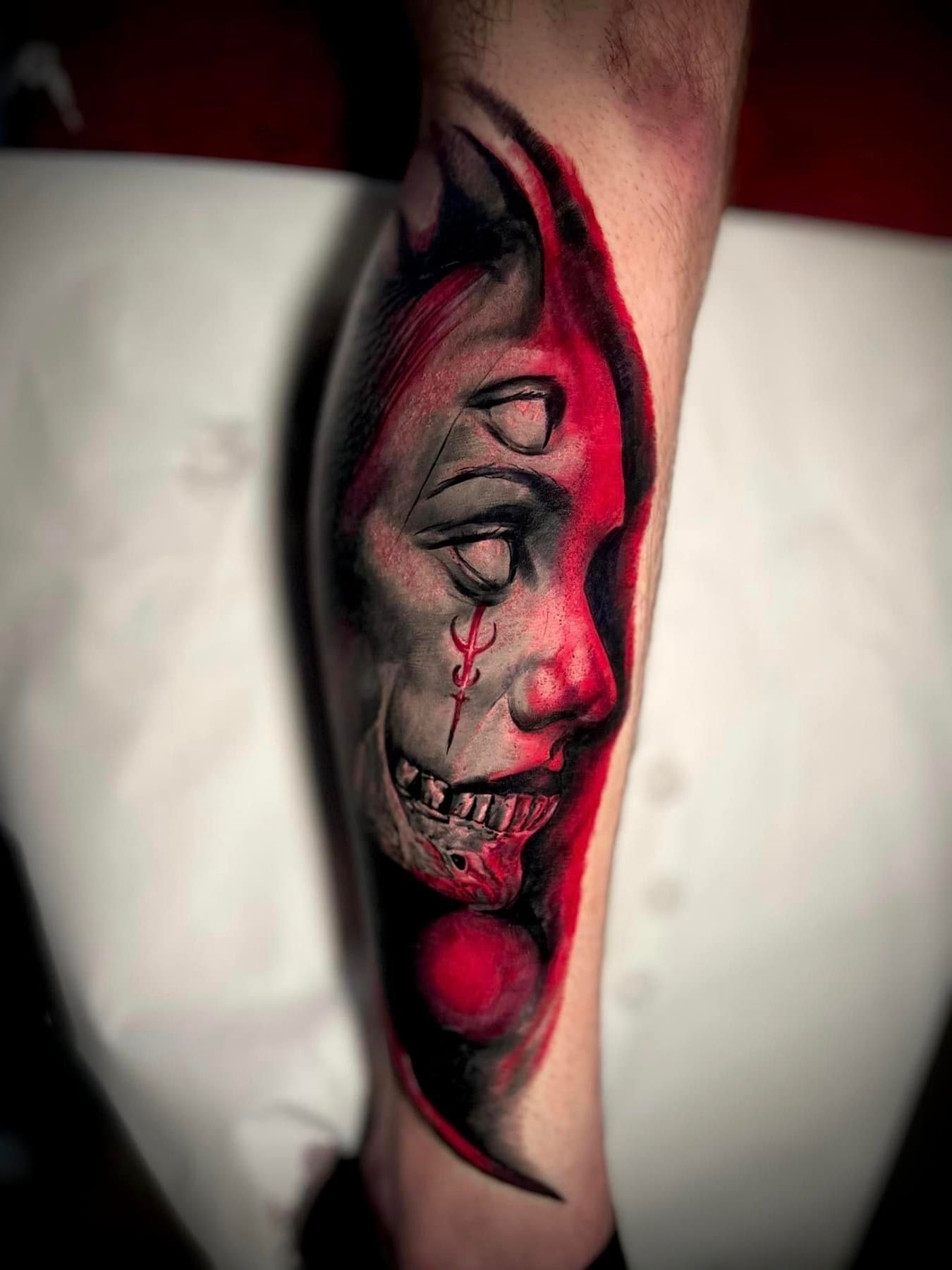 Tattoo of a grotesque face on a leg, with a red background. One eye closed, and mouth open showing teeth.