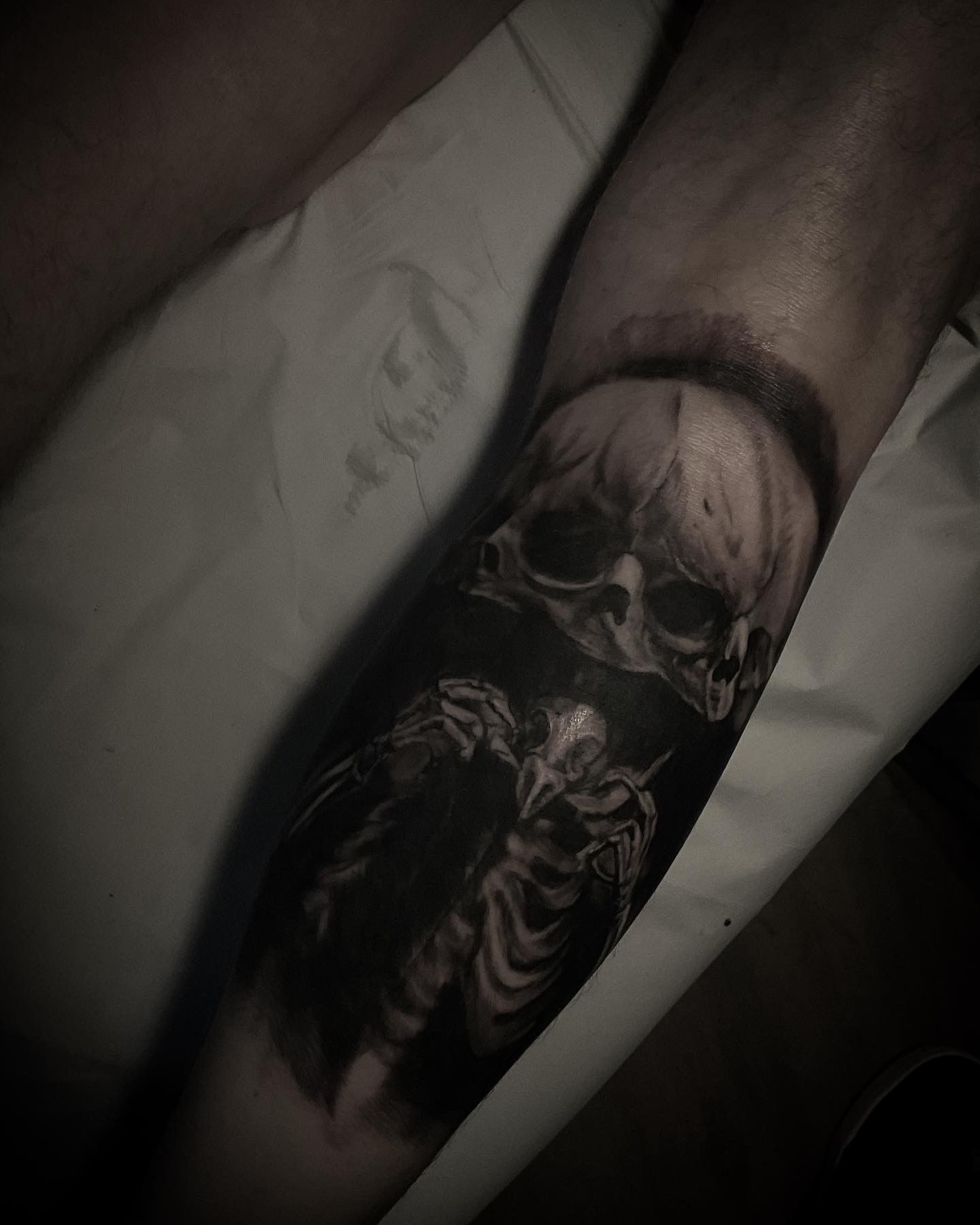 A black and white tattoo of a skull above a skeletal figure, on a human leg.