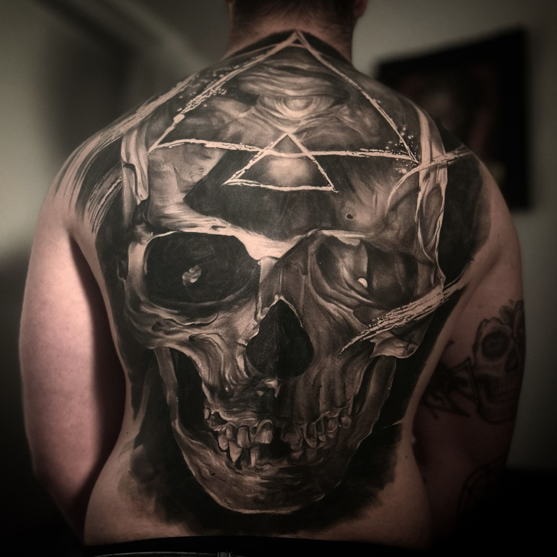 Back tattoo of a detailed skull with geometric shapes. Black and gray shading with visible teeth and eye sockets.