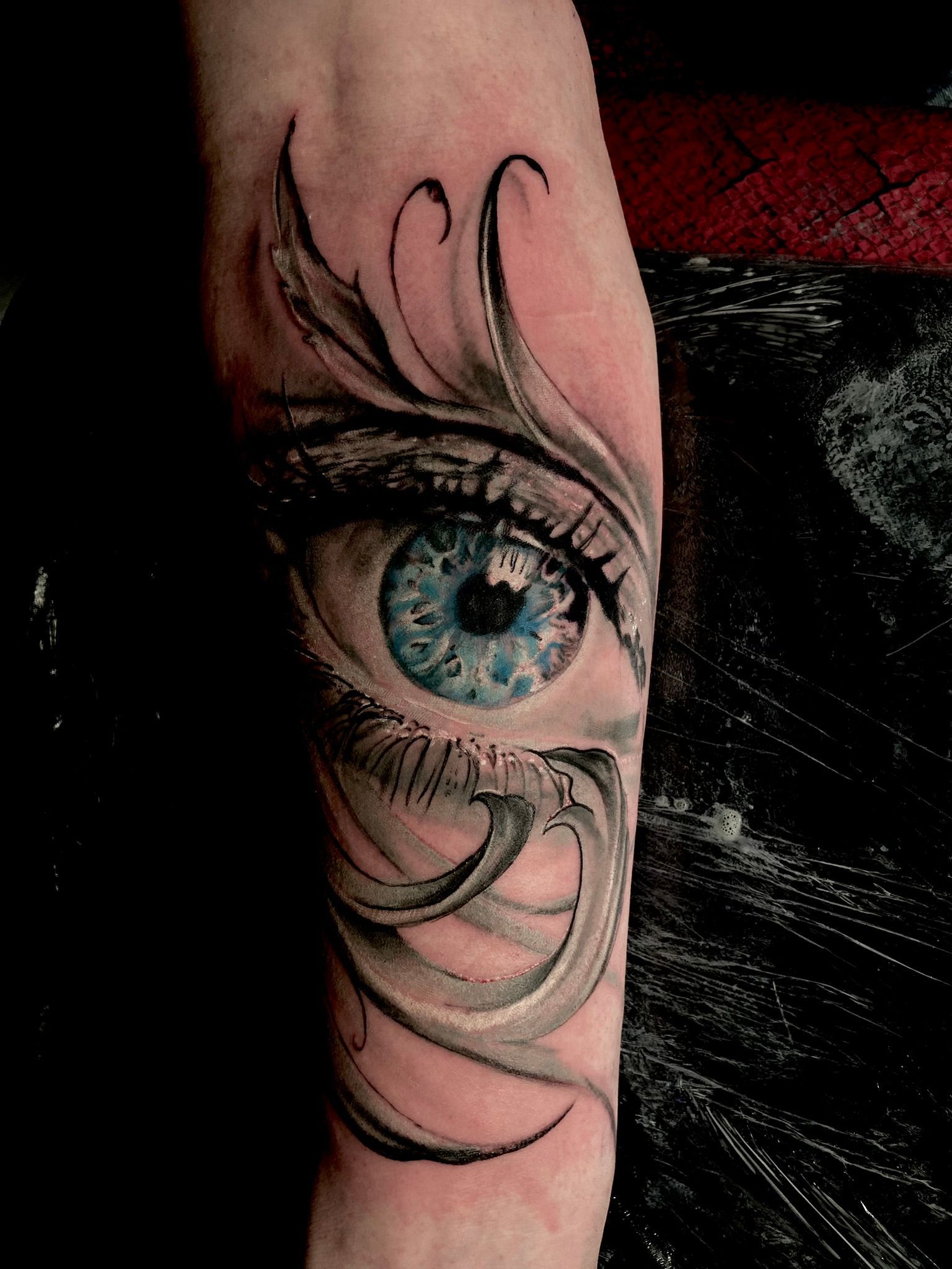 Tattoo of a blue eye on an arm, with stylized grey and black swirls around it.