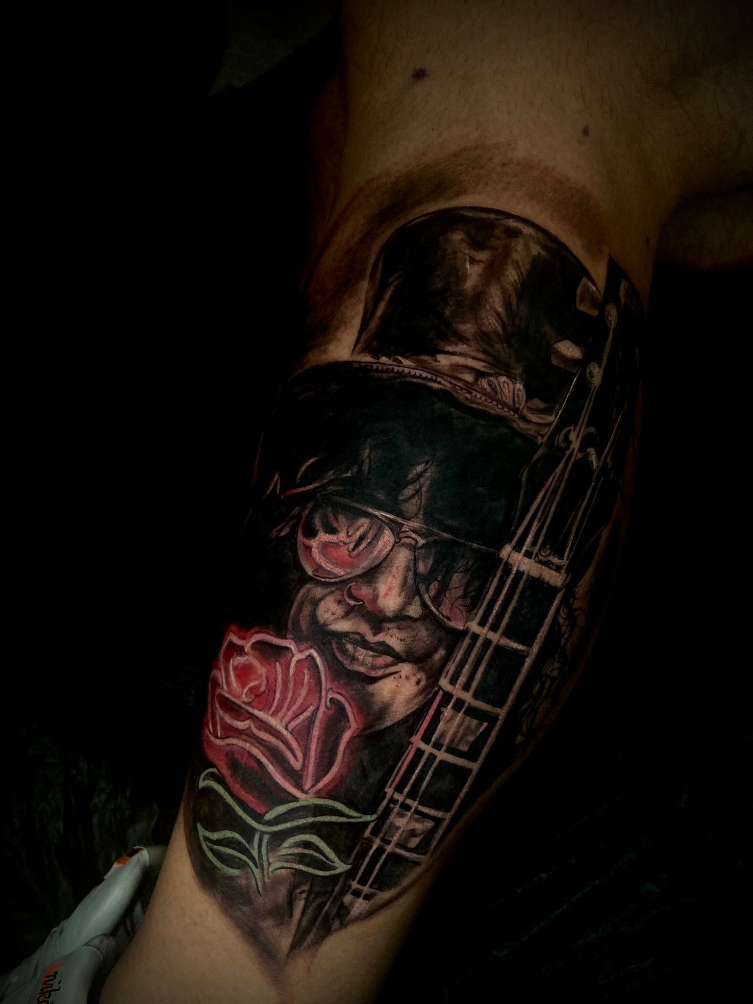 Tattoo on arm depicting a person wearing a hat and glasses, with a red rose and a guitar.