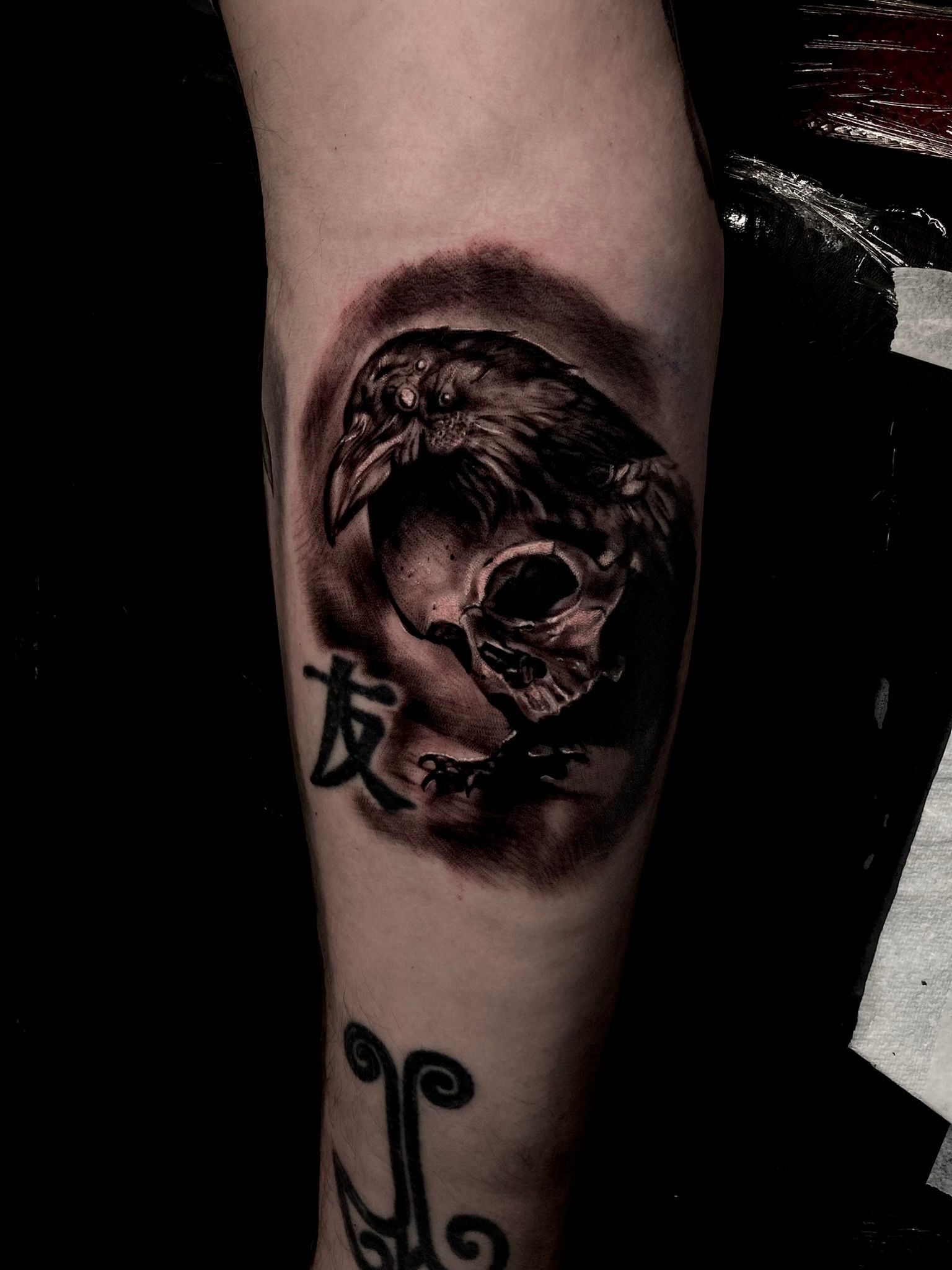 Tattoo of a raven perched on a skull; surrounded by dark shading. Chinese symbol 
