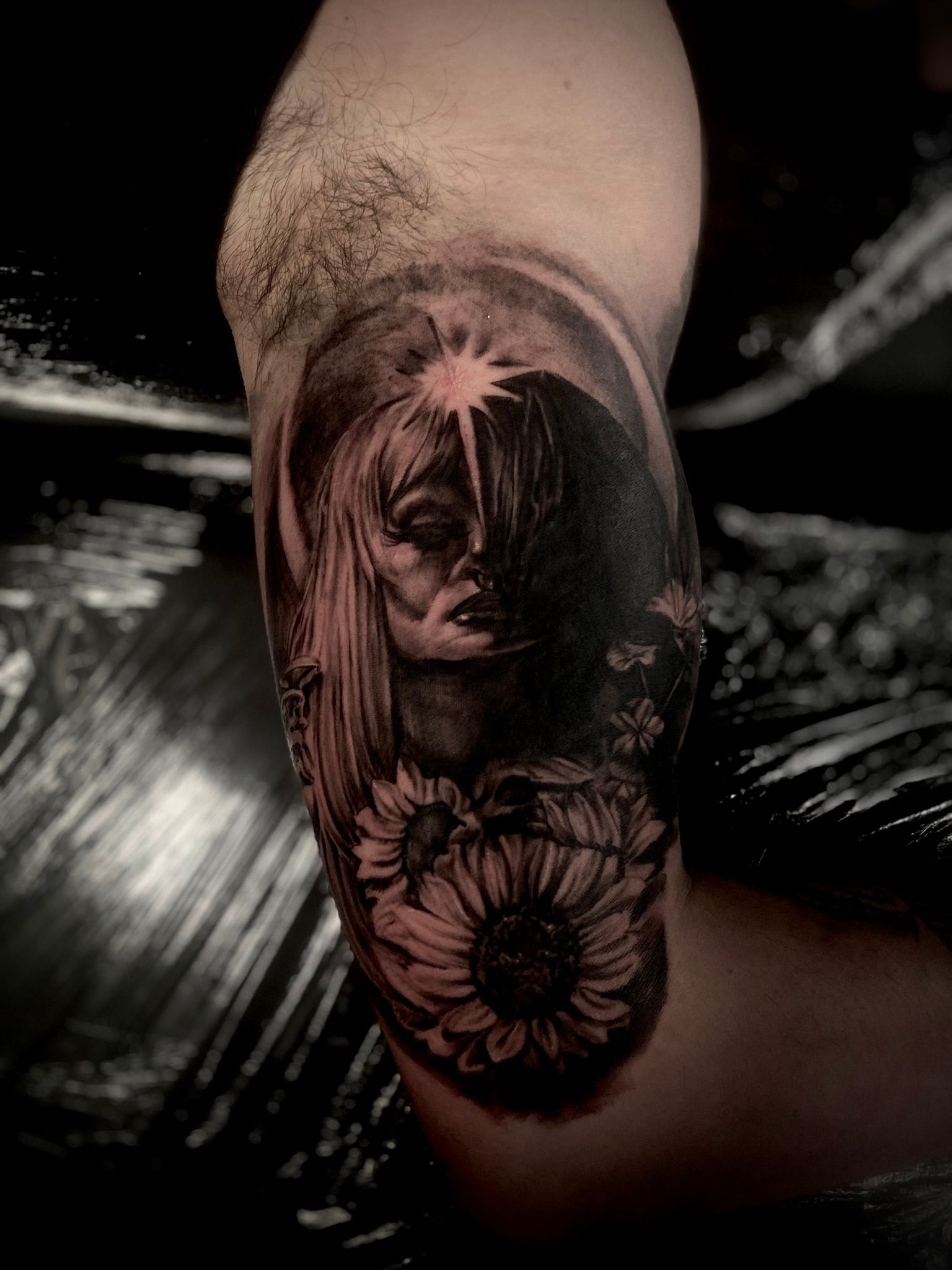 Black and gray tattoo of a woman with a half-lit face and sunflowers on an arm.