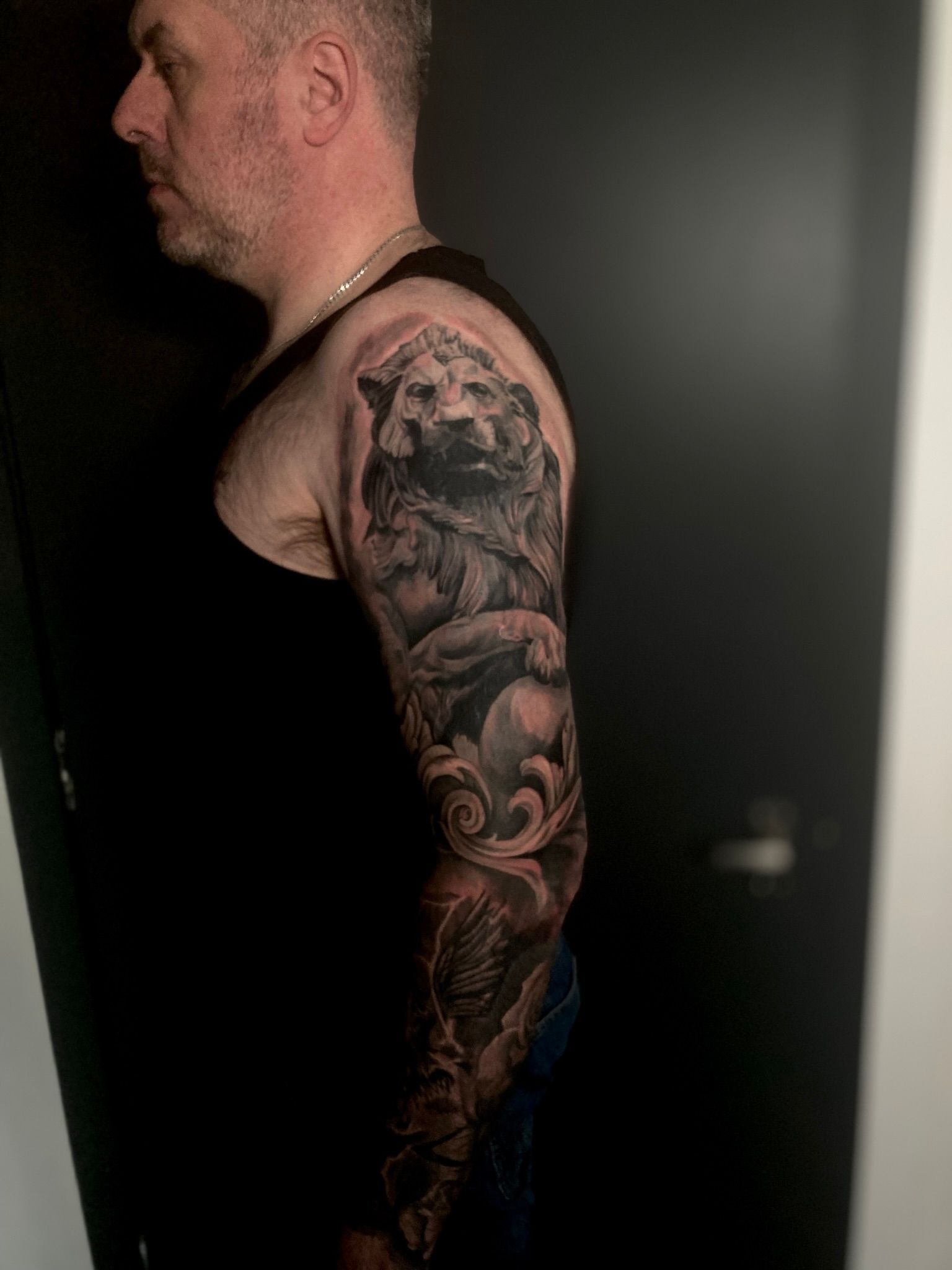 Man with full sleeve tattoo featuring a lion statue design, in a dark room.