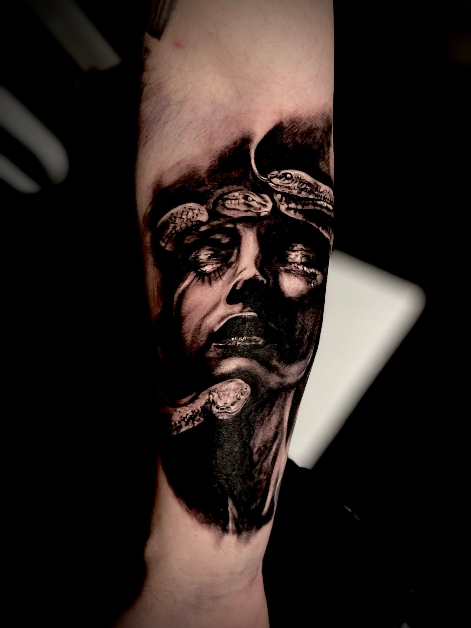 Black and white tattoo of a person’s face, partially obscured by shadows, with snake-like elements.