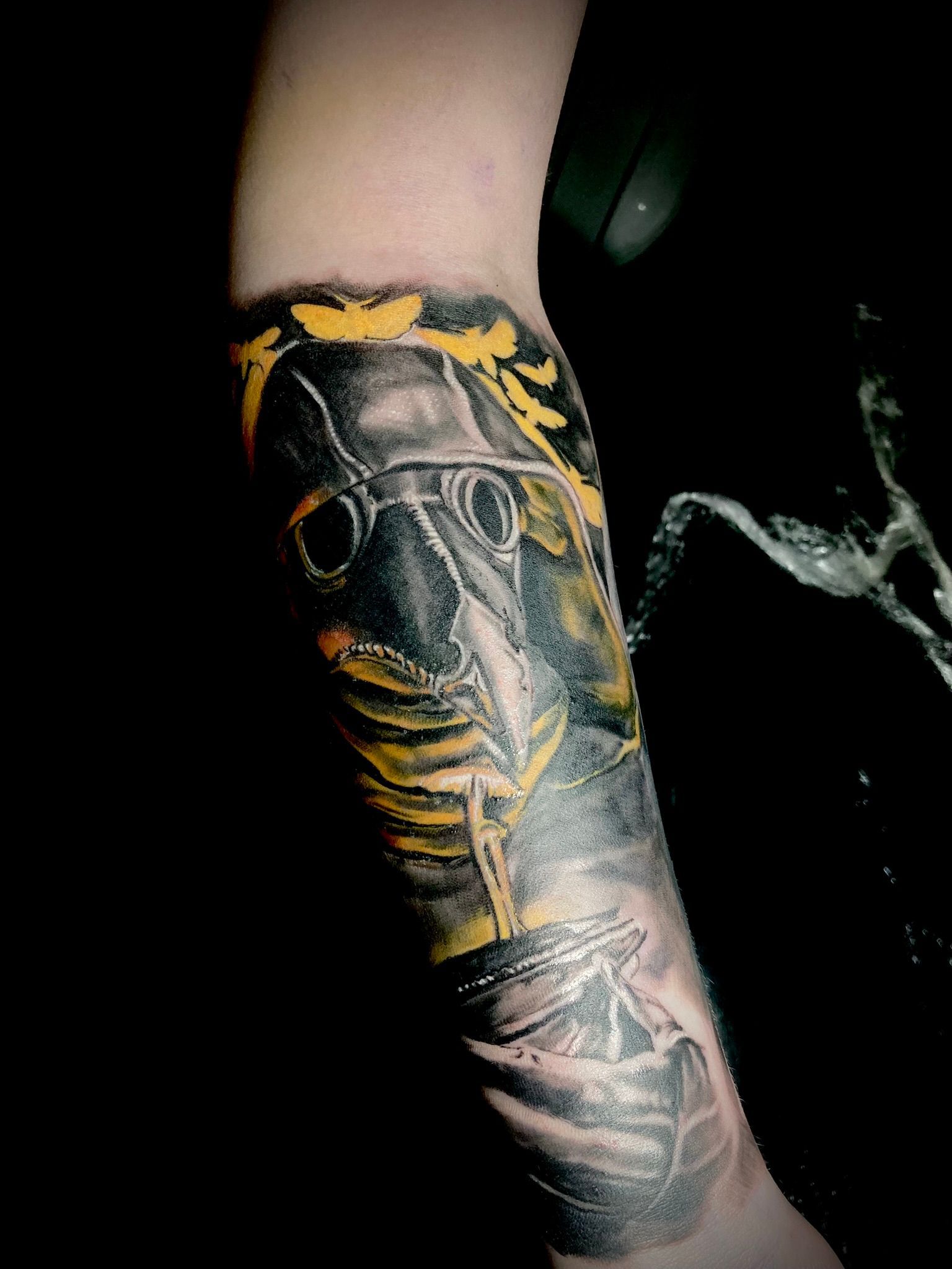 Tattoo of a plague doctor with a yellow mask and bird-like beak on a forearm. Black and grey shading.