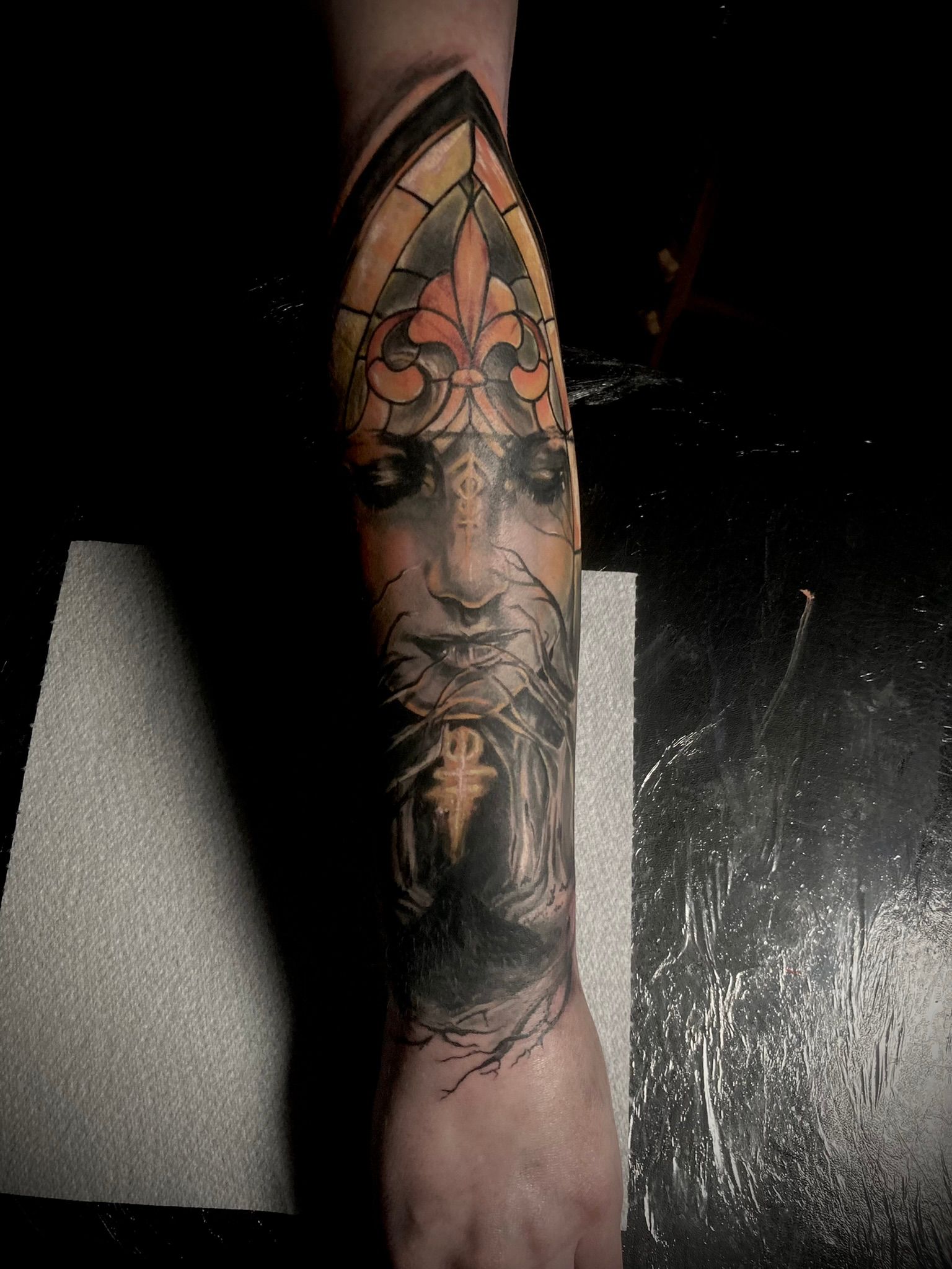 Detailed tattoo on an arm, featuring a face with a fleur-de-lis and stained glass window design in yellow and black.