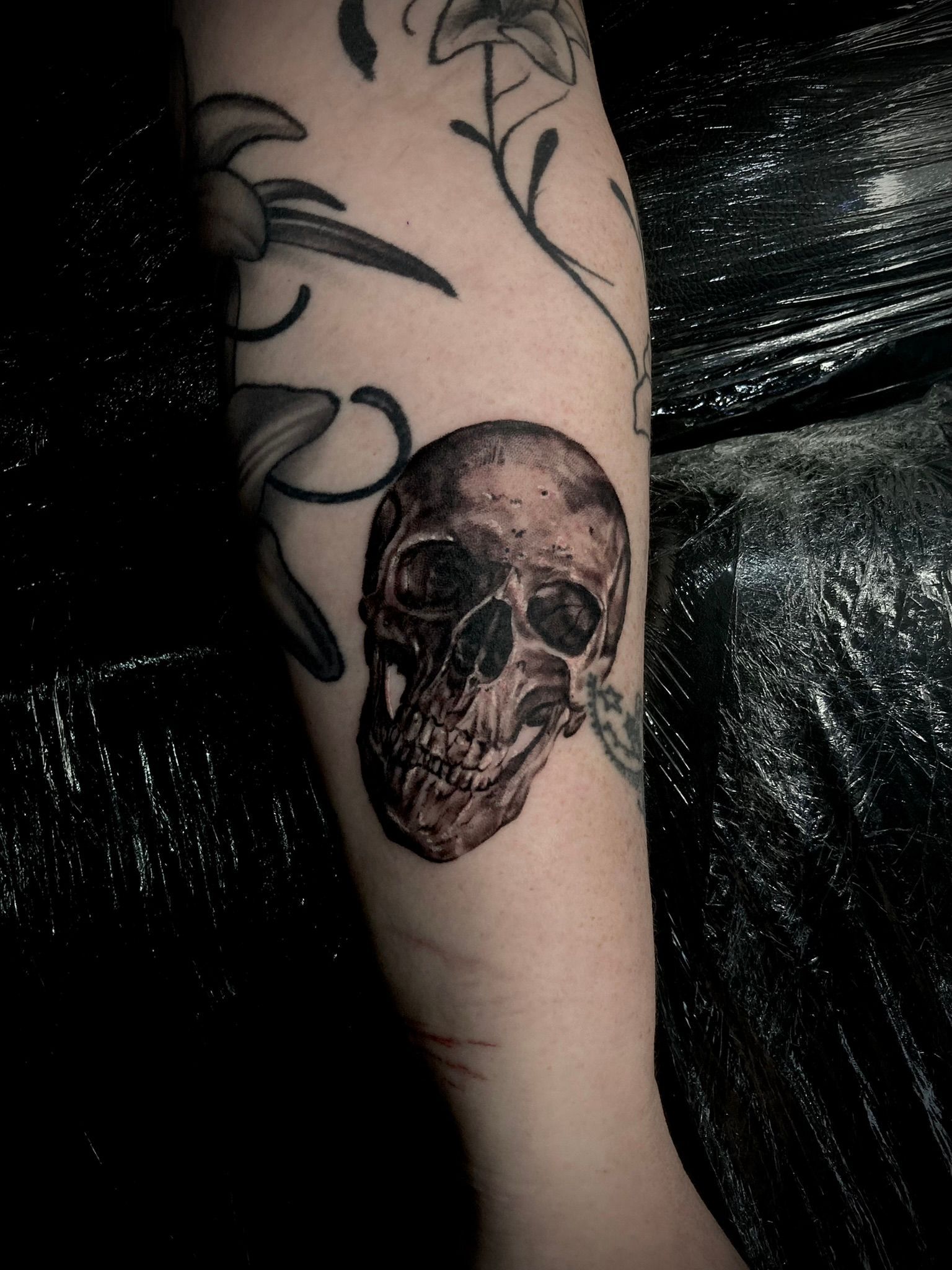 Tattoo of a skull on an arm, with floral designs partially visible. Dark, detailed shading.