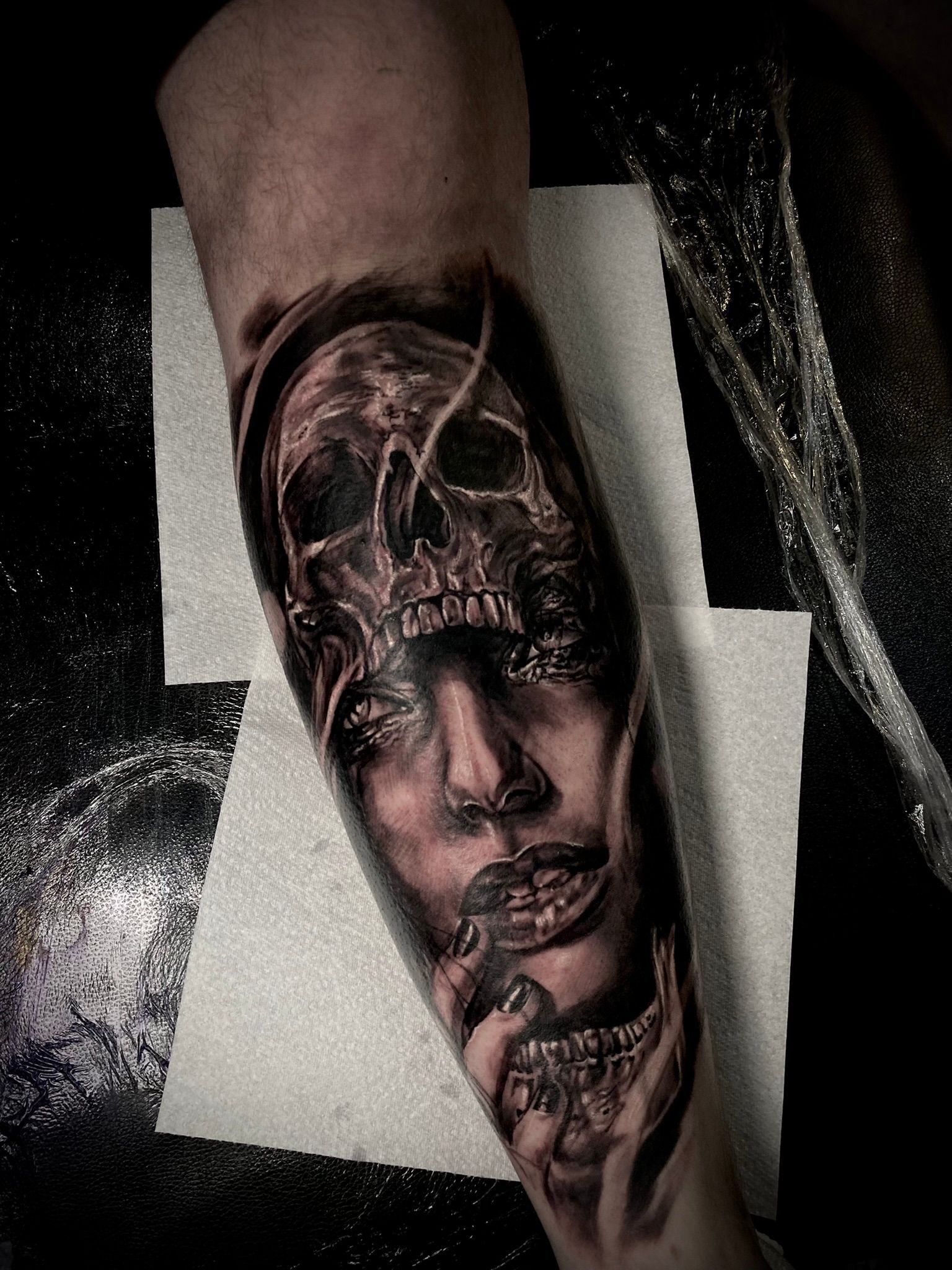 Tattoo of a skull merging with a woman's face on an arm, in black and gray ink.