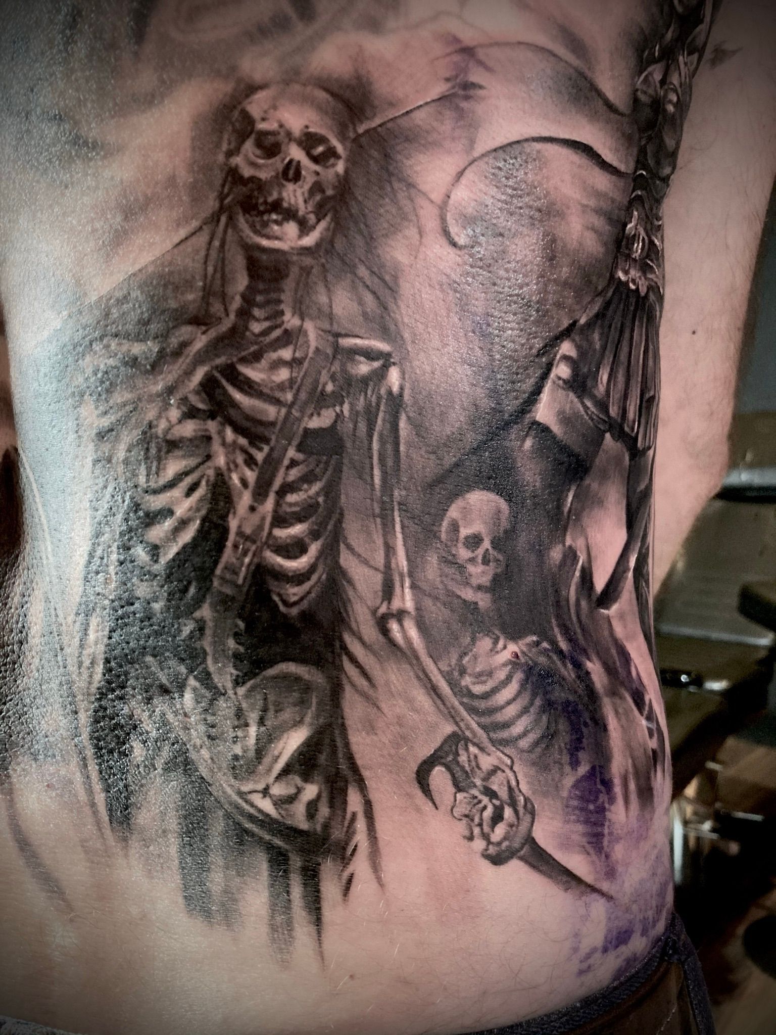 Black and gray skeletal tattoo on a torso. Figures hold weapons, shrouded in shadow.