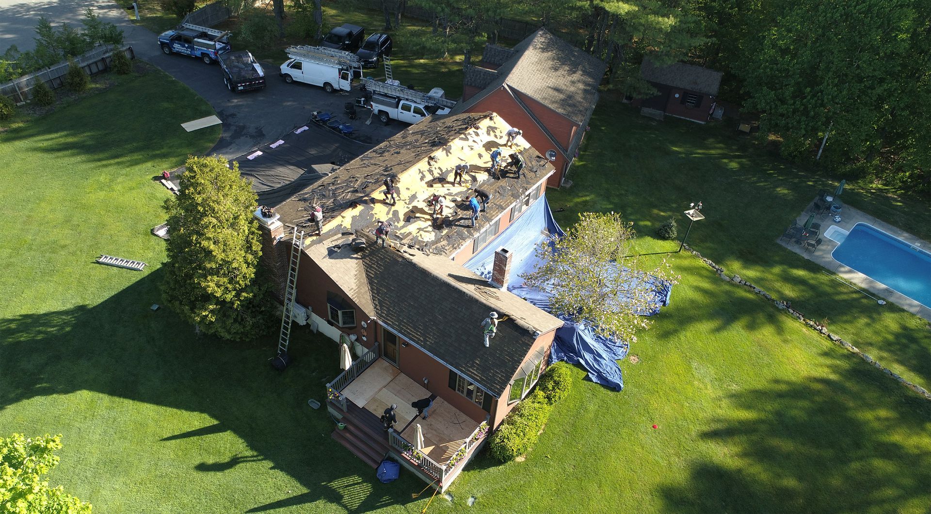 Aerial view of workers replacing a house roof, with a green lawn and trees in the background