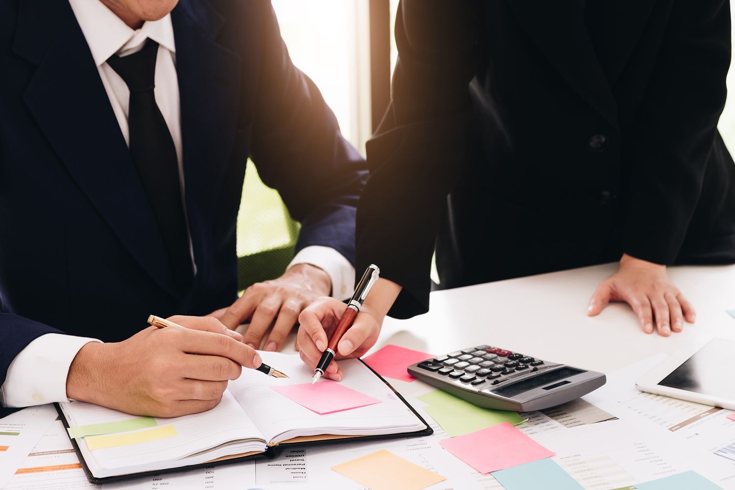 Two people in suits reviewing documents at a desk with a calculator and sticky notes.