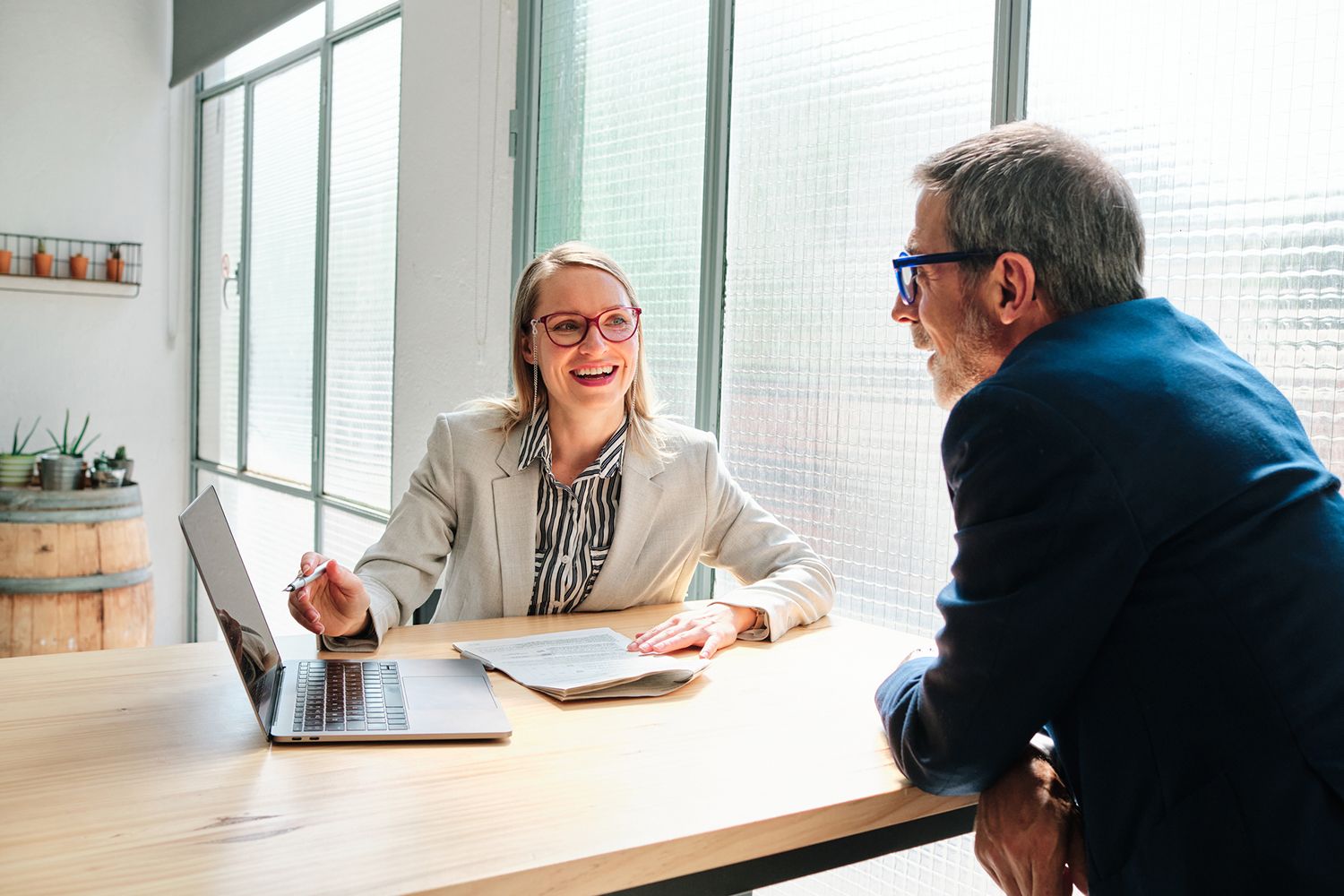 Woman in glasses smiles, working with a man at a table with a laptop in an office.