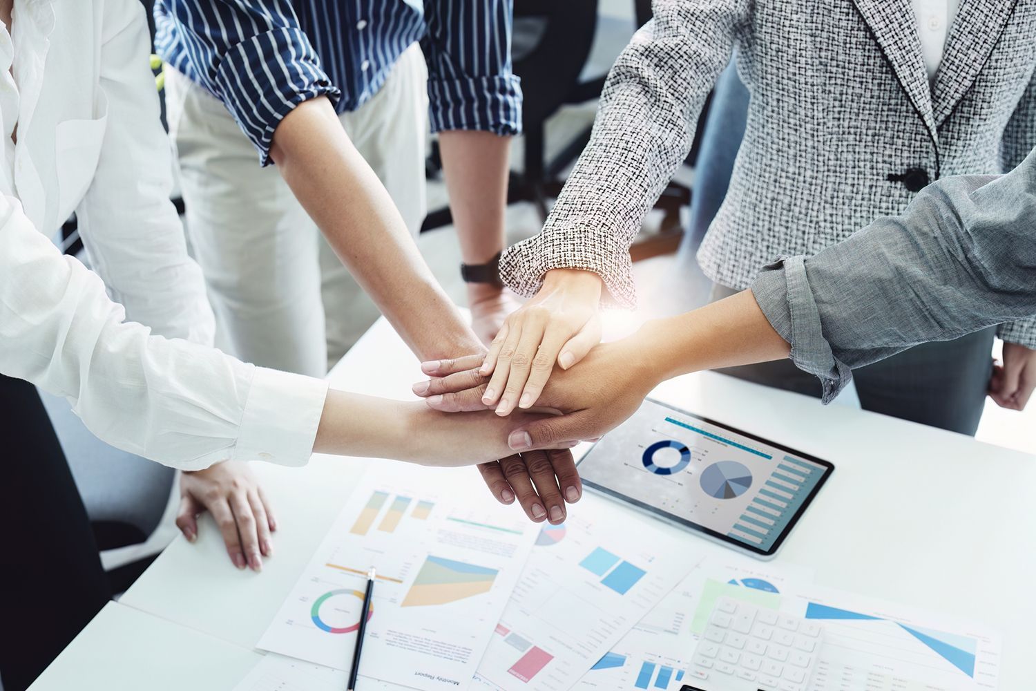 Hands stacked in a team huddle over charts and a tablet on a desk.