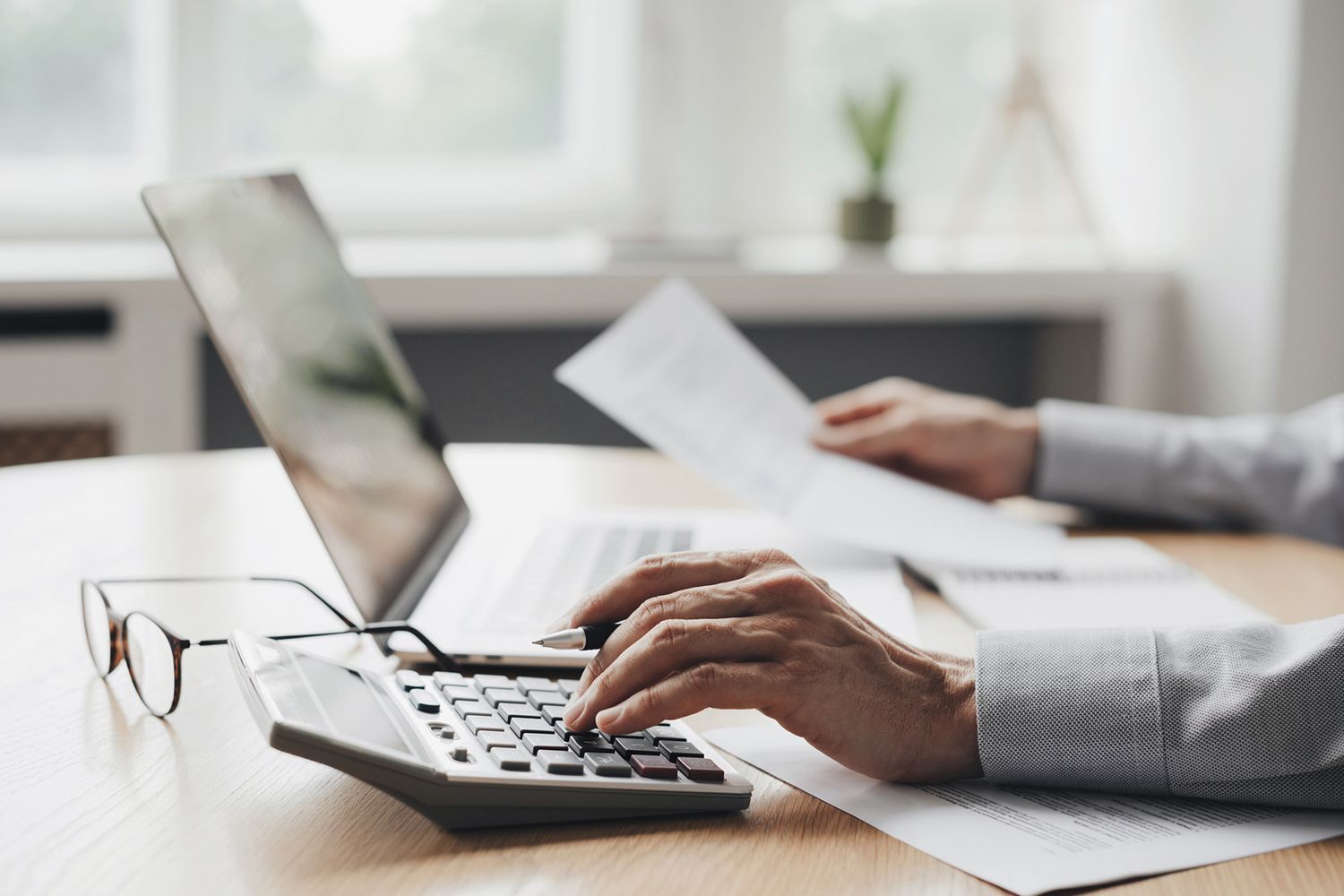 Person using a calculator and laptop at a desk, reviewing papers in a well-lit office setting.