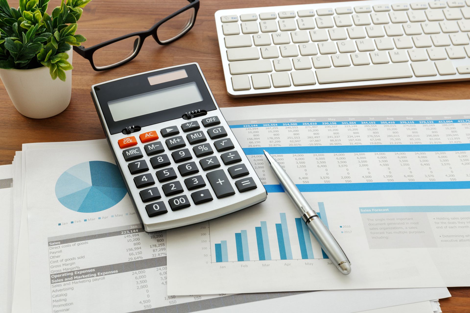 Calculator and pen on financial reports beside keyboard and glasses on a desk