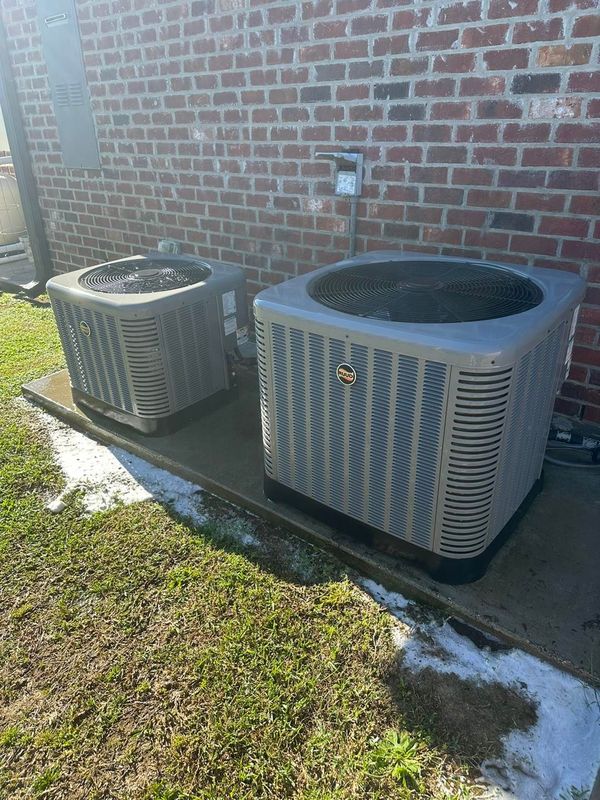 Two air conditioning units side-by-side on concrete, against a red brick wall, and green grass.