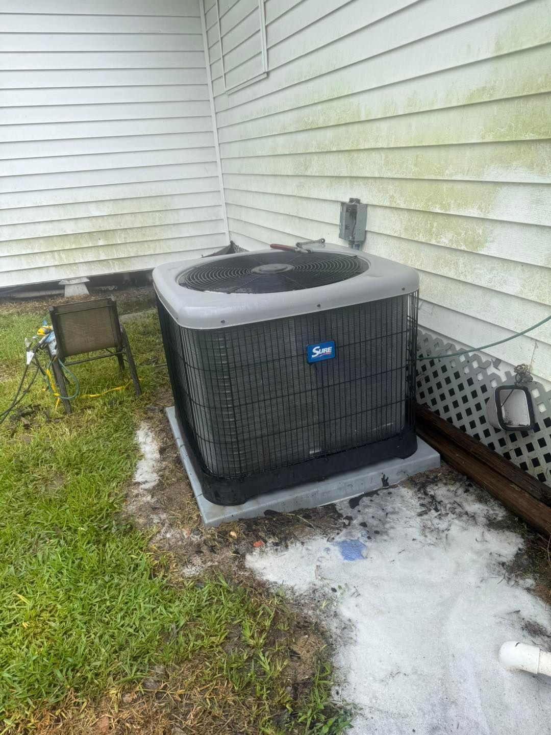 Air conditioner unit outside a building, surrounded by grass and a white wall with mold.