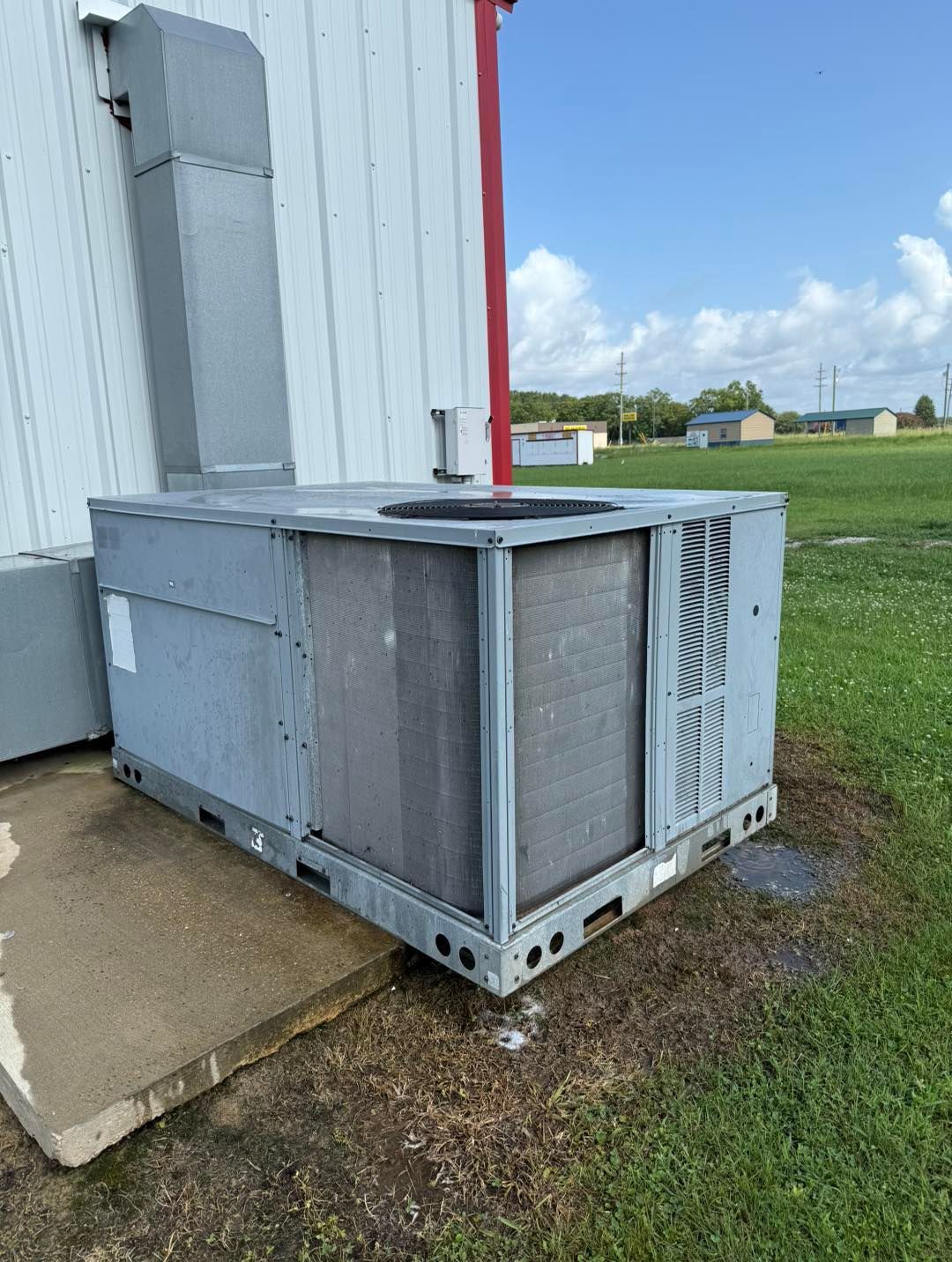 An industrial air conditioning unit on a concrete pad next to a white building with a red support beam.
