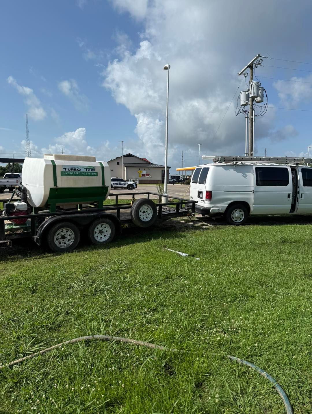 White van towing a trailer with a water tank on grass near a power pole under a cloudy sky.