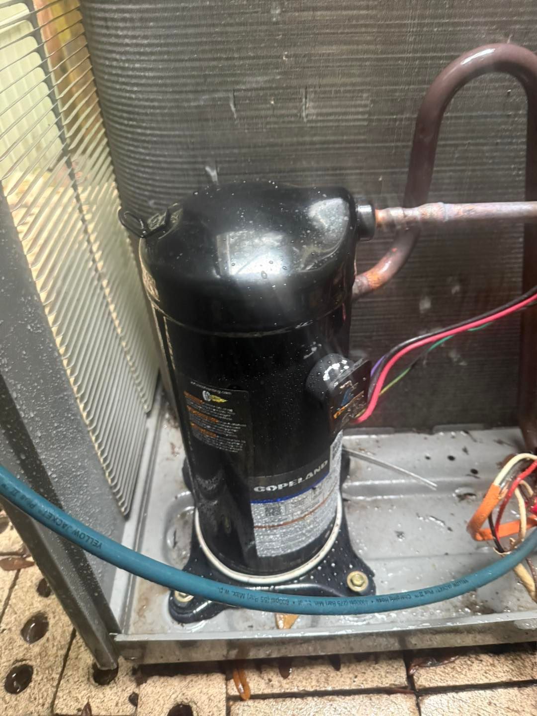 Air conditioner compressor in outdoor unit; black cylinder, copper piping, electrical wires.