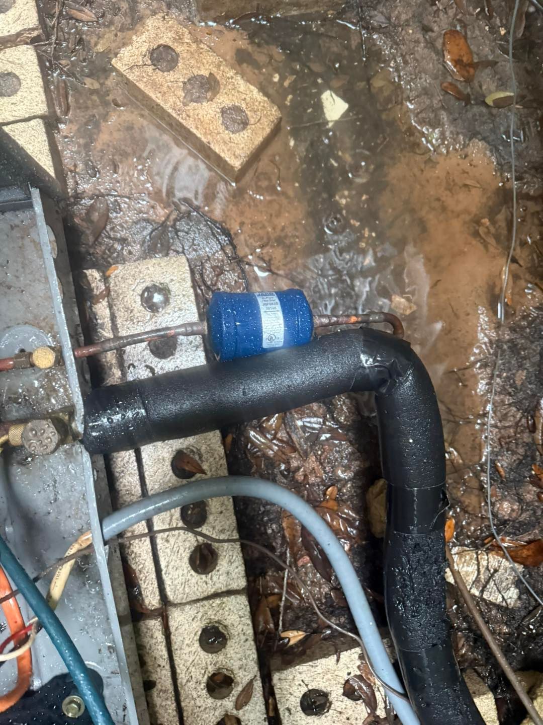 Outdoor air conditioning unit with a blue filter and black insulated pipes, in a flooded area.