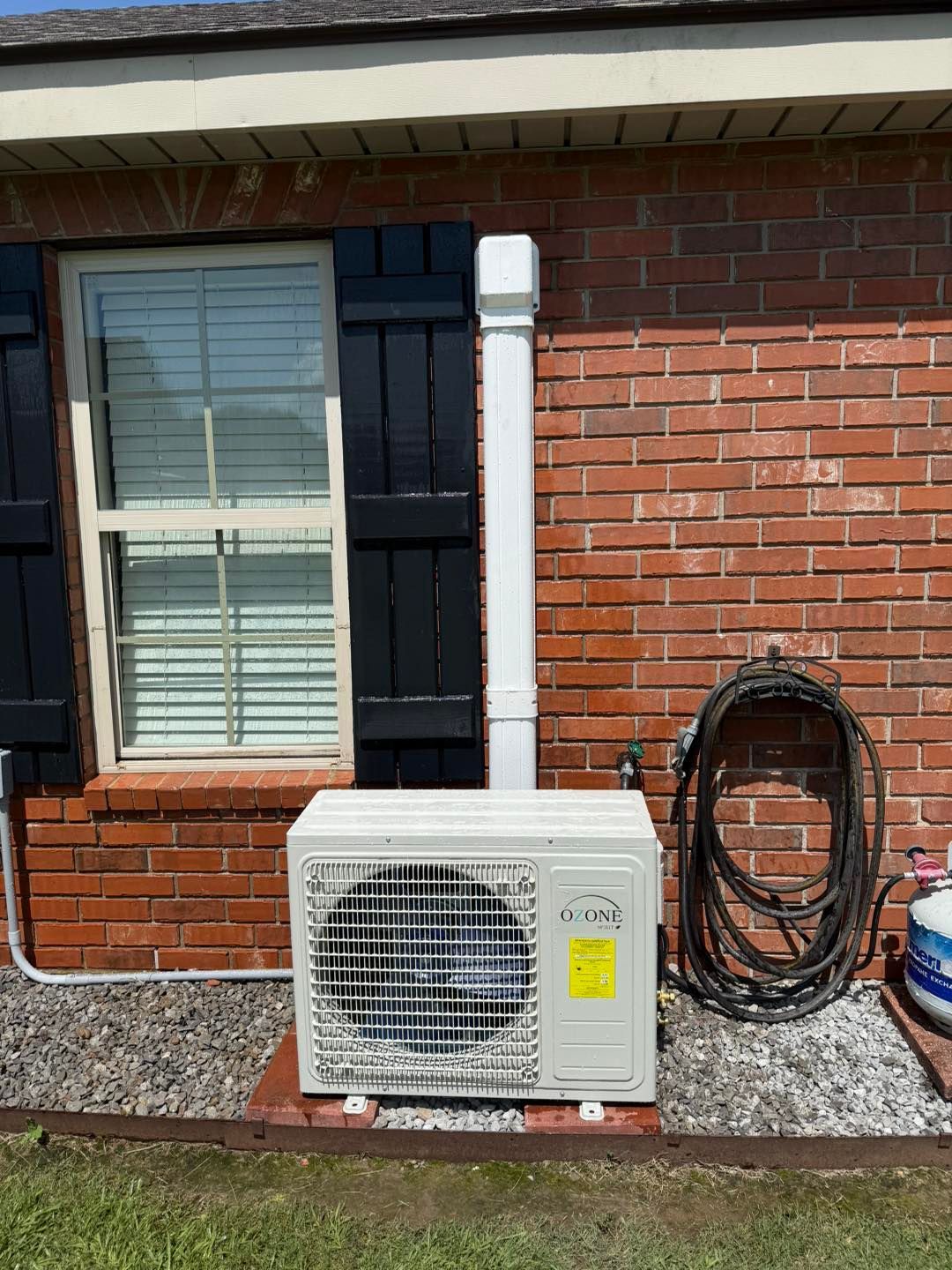 Exterior air conditioning unit next to a brick building and a window with black shutters.