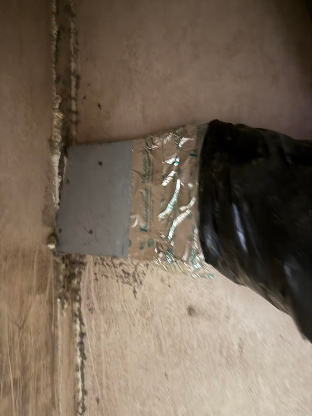 Black-gloved hand applying sealant to a silver tape patch on wood. Vertical wood joint is visible.