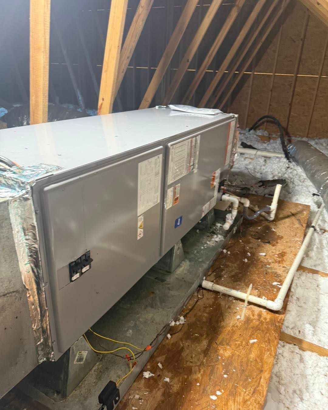 HVAC unit in an attic. Gray metal rectangular unit with white pipes and wooden beams in the background.