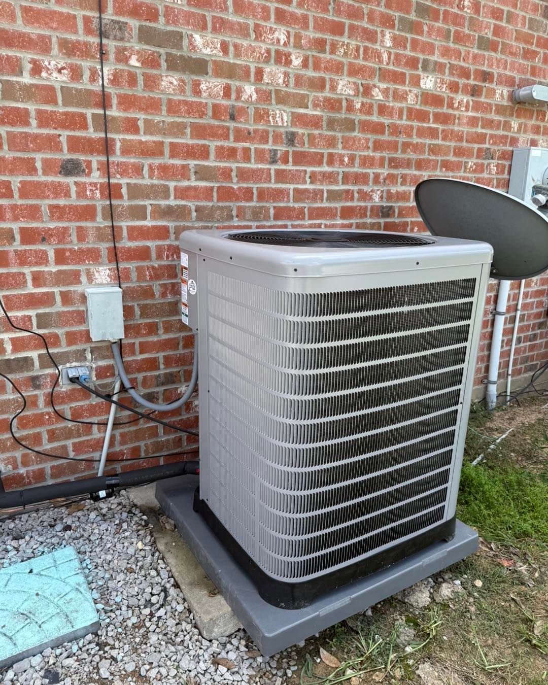 Air conditioning unit against a brick wall. Gray unit sits on a gray platform. Electrical box and satellite dish nearby.