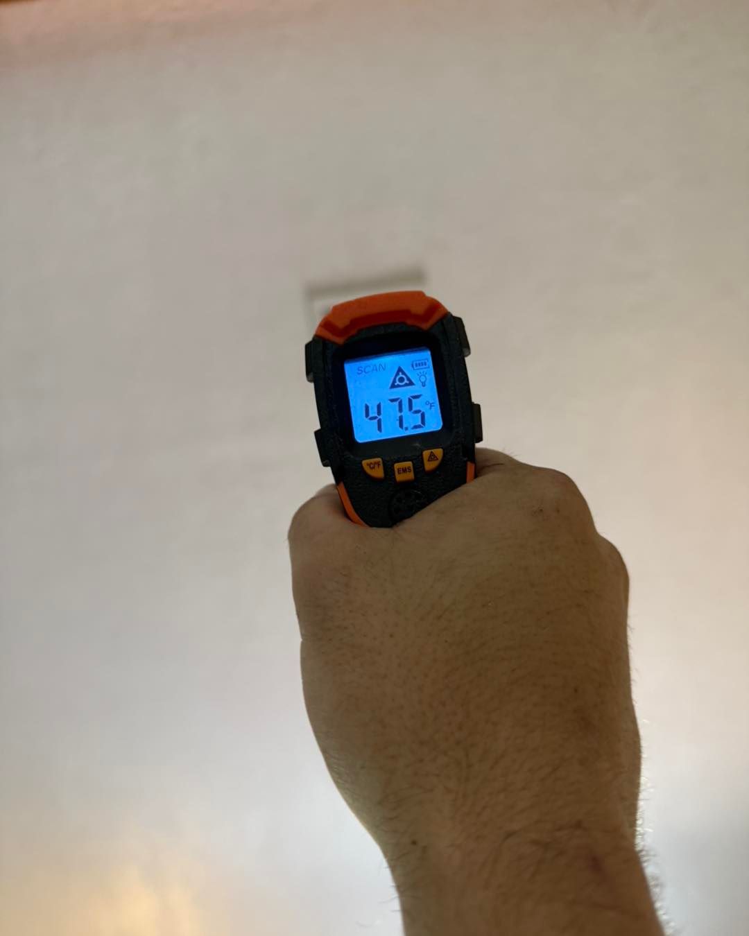 A hand holding a thermal gun measures a white ceiling, displaying a temperature of 47.5 degrees.