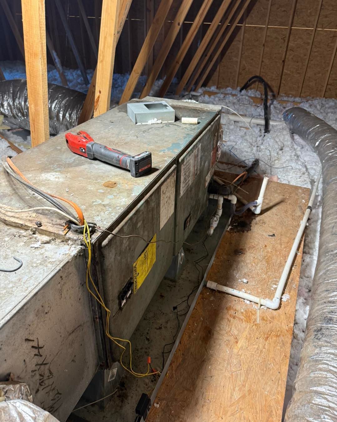 HVAC unit in an attic. Dirty, gray metal box with electrical wiring, insulation, and ductwork.  A flashlight and gray box are on top.